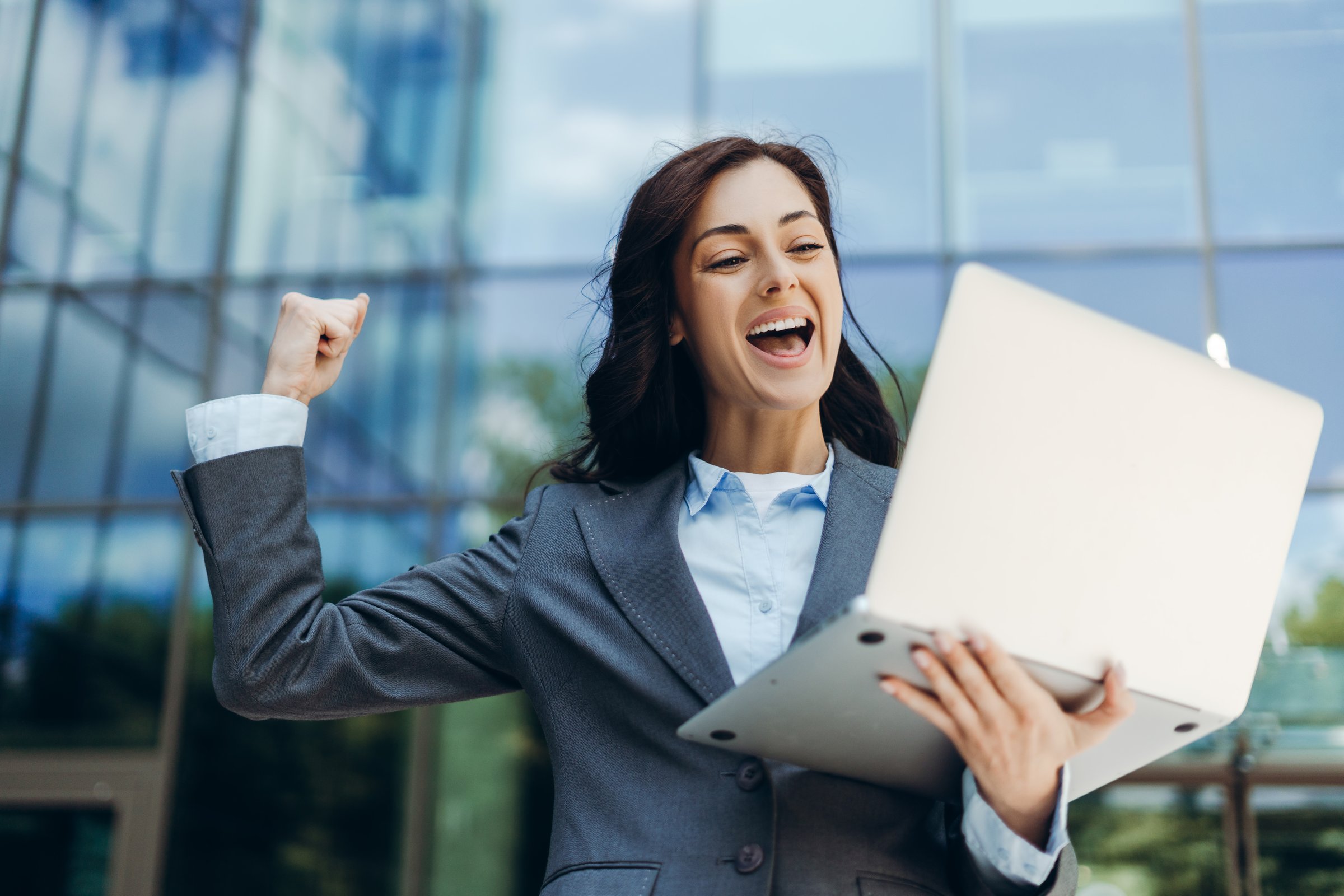 Enthusiastic businesswoman in a suit is raising her fist in celebration while holding a laptop, expressing joy and achievement in front of a modern office building. Online technology concept