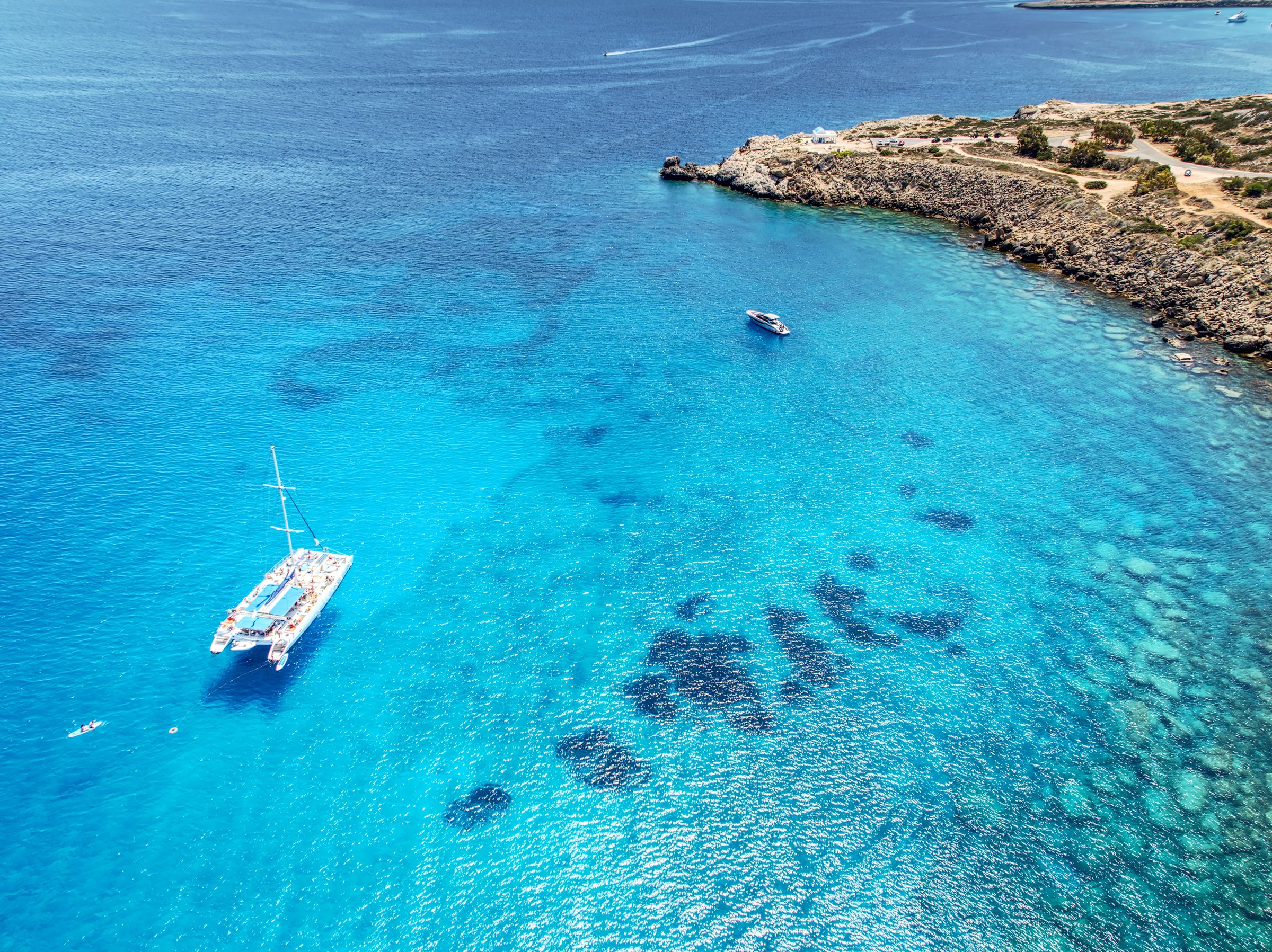 Aerial view of a catamaran and kayak in clear blue waters near a rocky coastline, offering a serene and picturesque seascape.