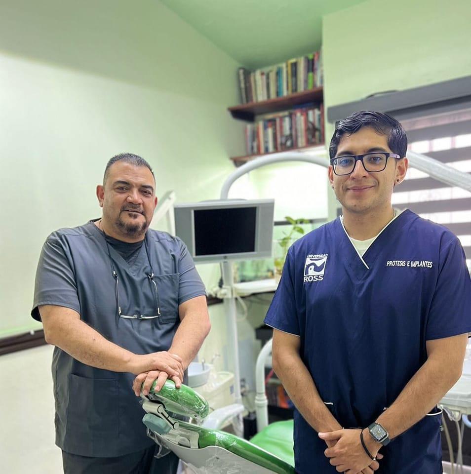 Two dentists standing in a dental office, one in gray scrubs and the other in blue. Dental equipment is visible around them.