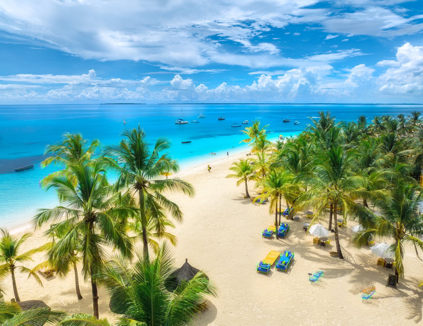 Aerial view of white sandy beach with palm trees, sunbeds, umbrellas, yachts, boats, blue ocean, sky with clouds at sunset. Summer in Kendwa, Zanzibar island. Tropical landscape. Clear sea. Top view