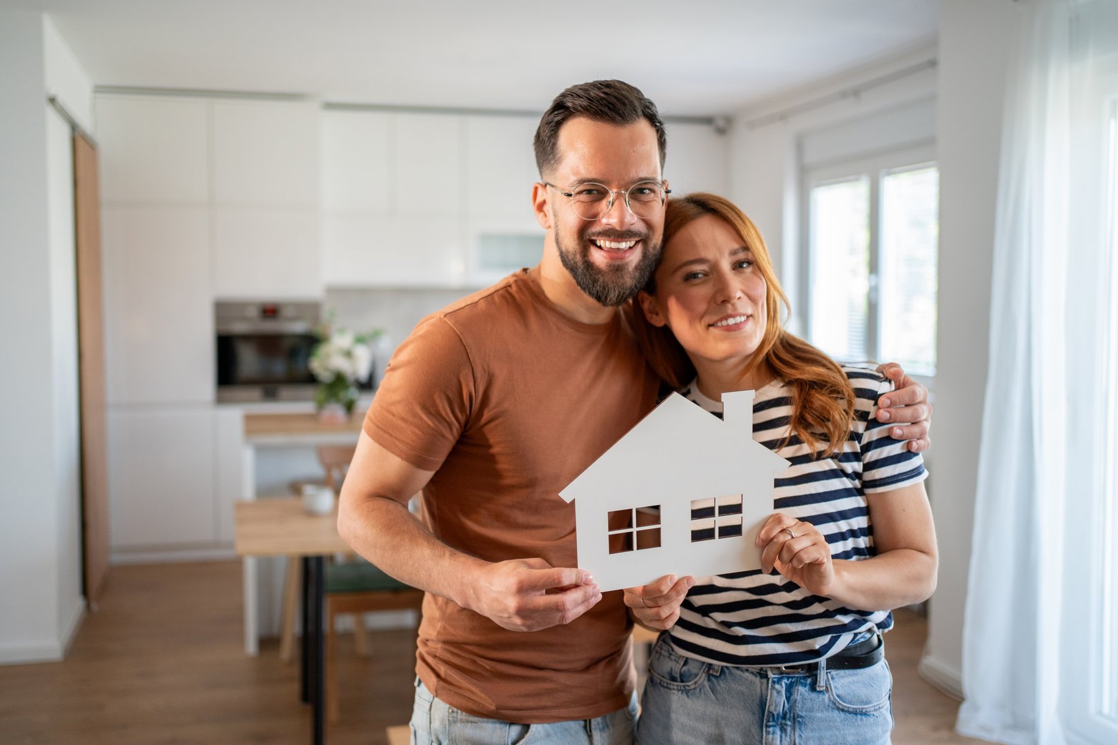 Happy couple embracing and holding a white paper house, smiling at the camera in their modern new home, celebrating buying their first house together