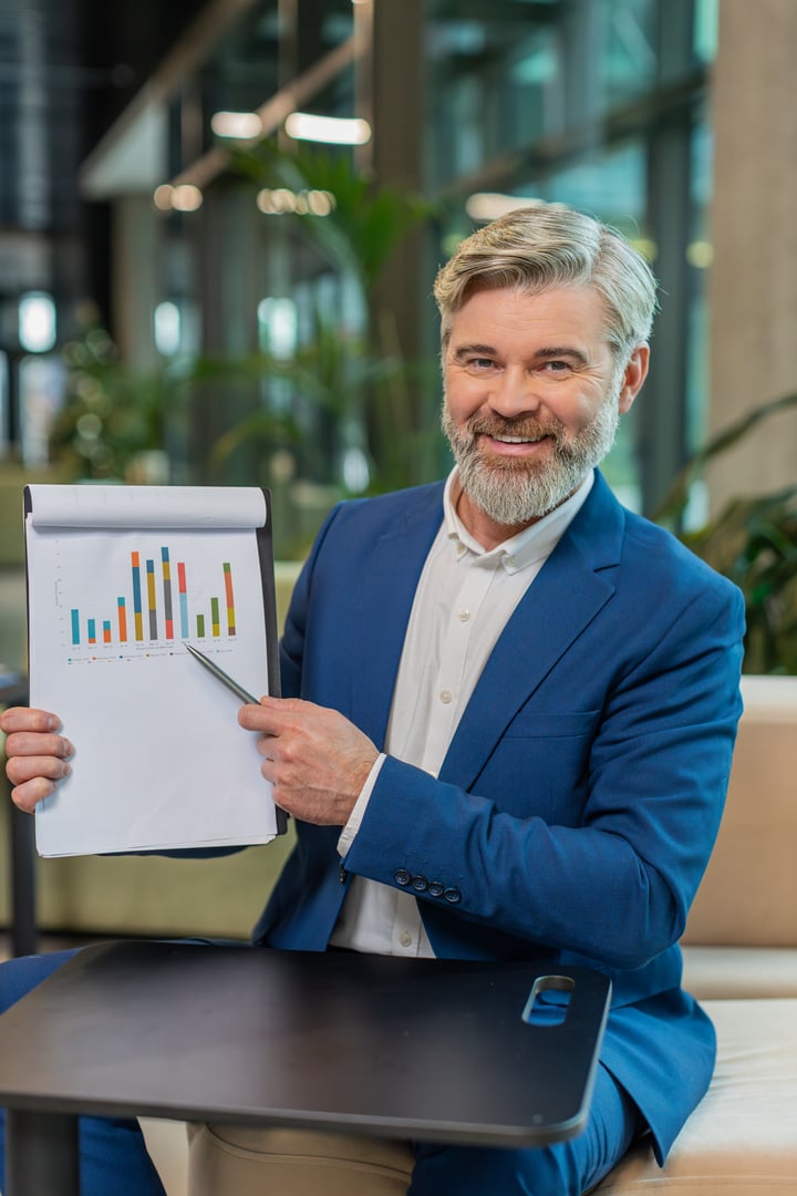 Mature Caucasian boss businessman making online presentation video call, demonstrates graphic analytics, planning budget, statistic. Handsome guy in suit at office lobby looking at camera, talking.