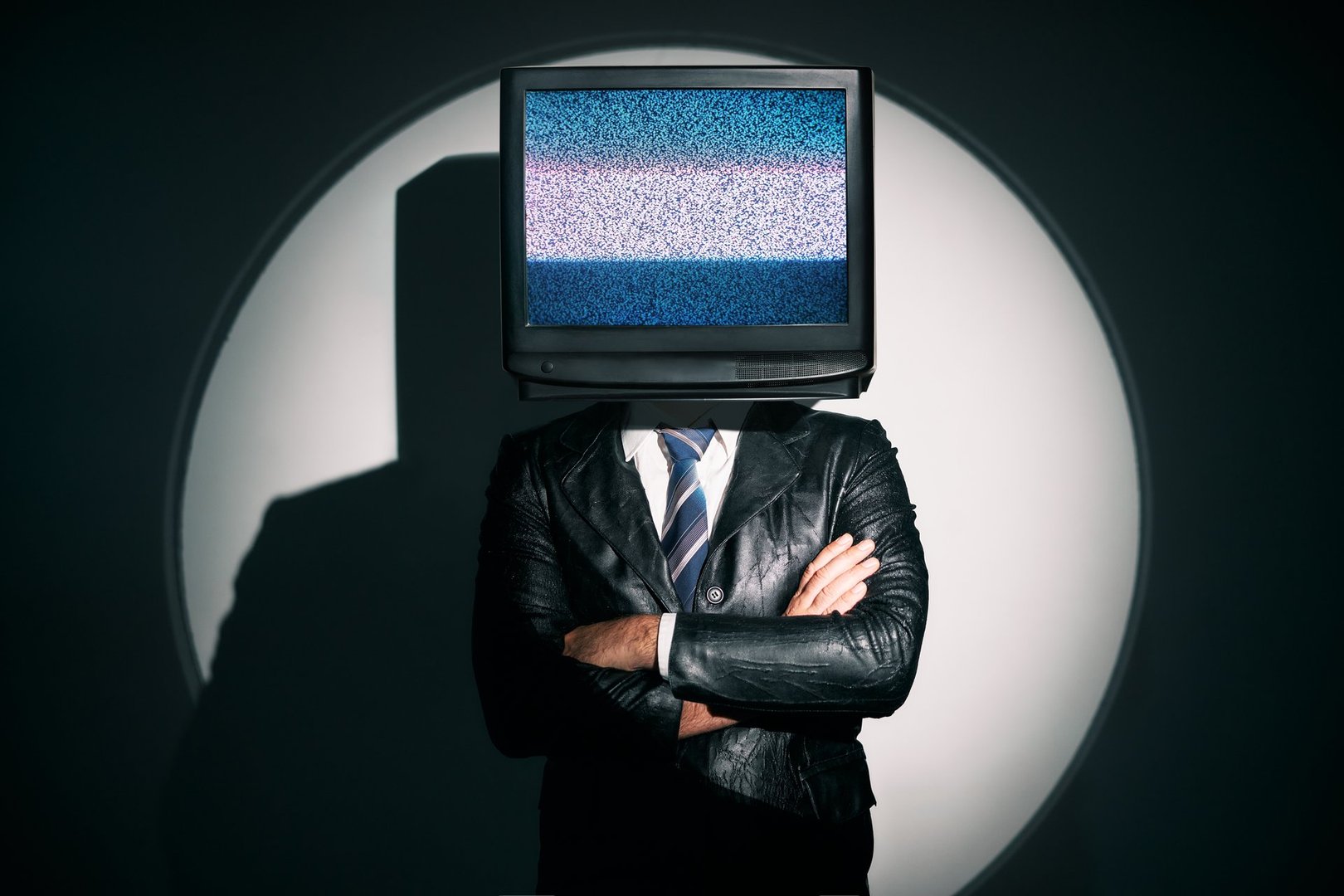 Man with TV instead of head. Media zombie concept with television addicted male in black coat with crossed arms posing on studio in the spotlight. Conceptual, contemporary art collage. Surrealism