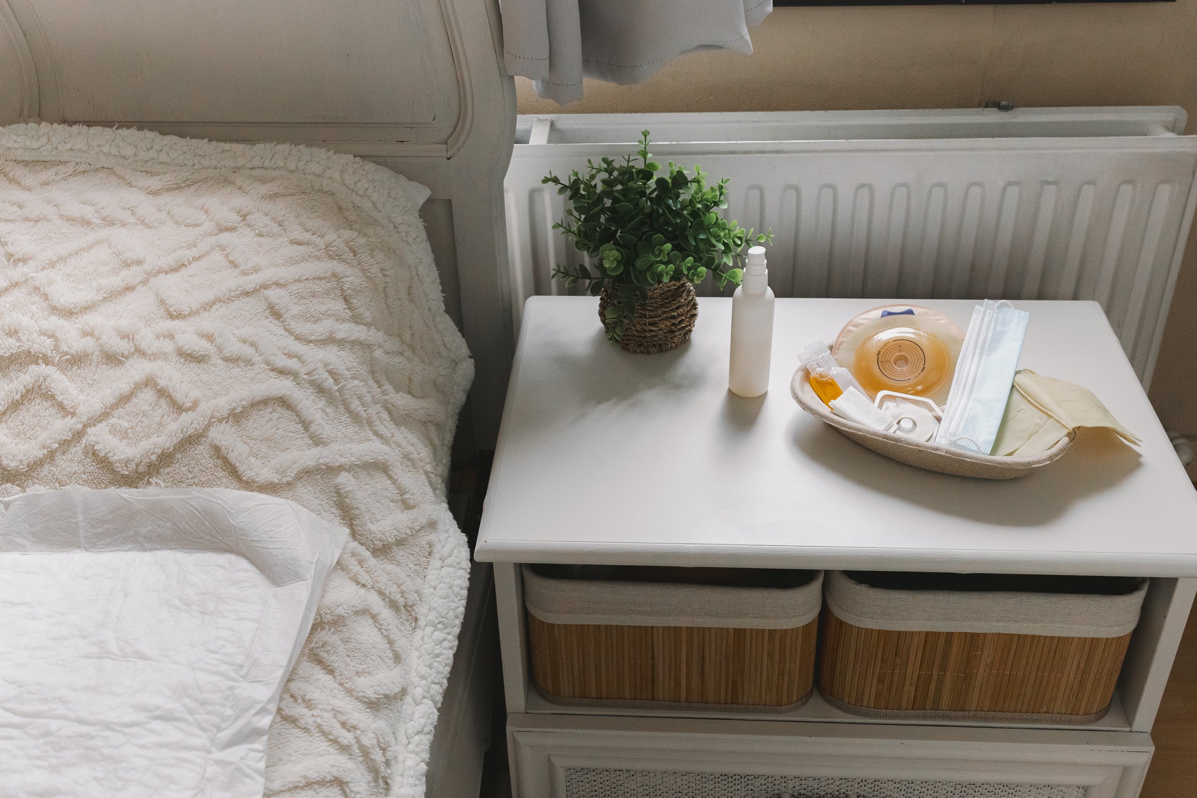 View of disposable cardboard tray with ostomy bag, mask, sterile gloves and medicines standing on nightstand with plant near bed during daytime in home clinic ward, side view medium close-up. Abdominal diseases concept, nurse services.