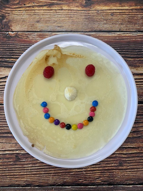 A smiling face made of candy and fruit on a pancake, with raspberries for eyes and a colorful candy mouth on a white plate.