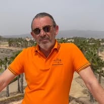 Man in an orange shirt and sunglasses standing outdoors with a desert landscape and palm trees in the background.