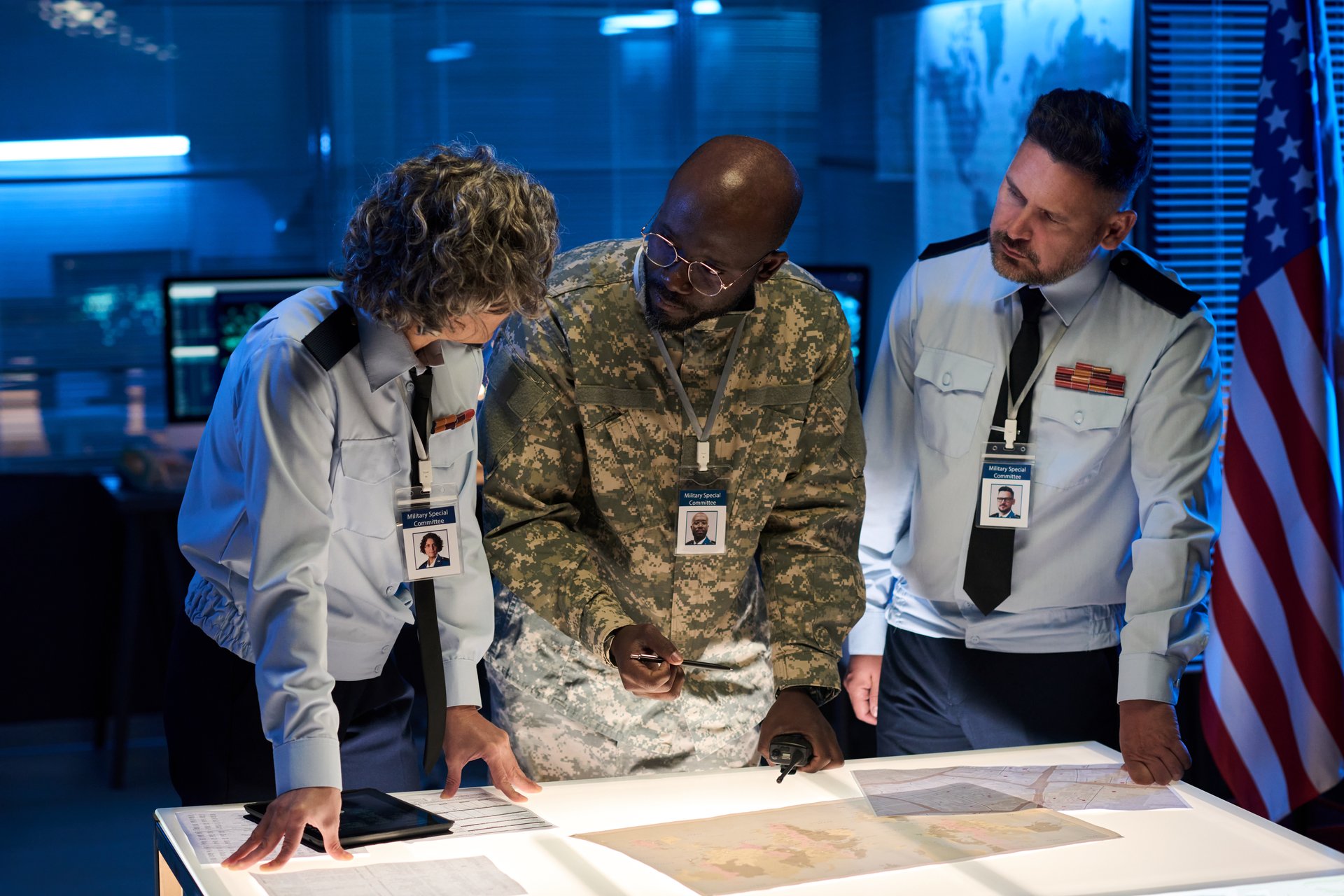 Group of three intercultural workers of military command center discussing map or other documents by table during meeting
