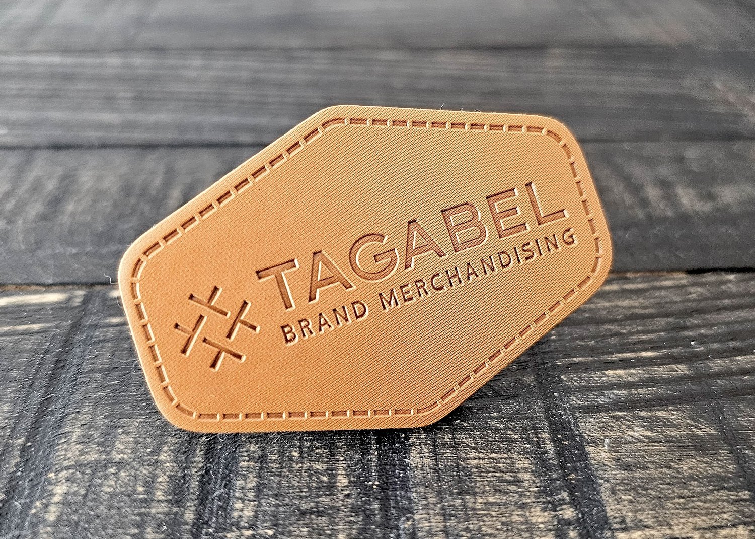 Leather patch with embossed TAGABEL Brand Merchandising text on a textured wooden surface.