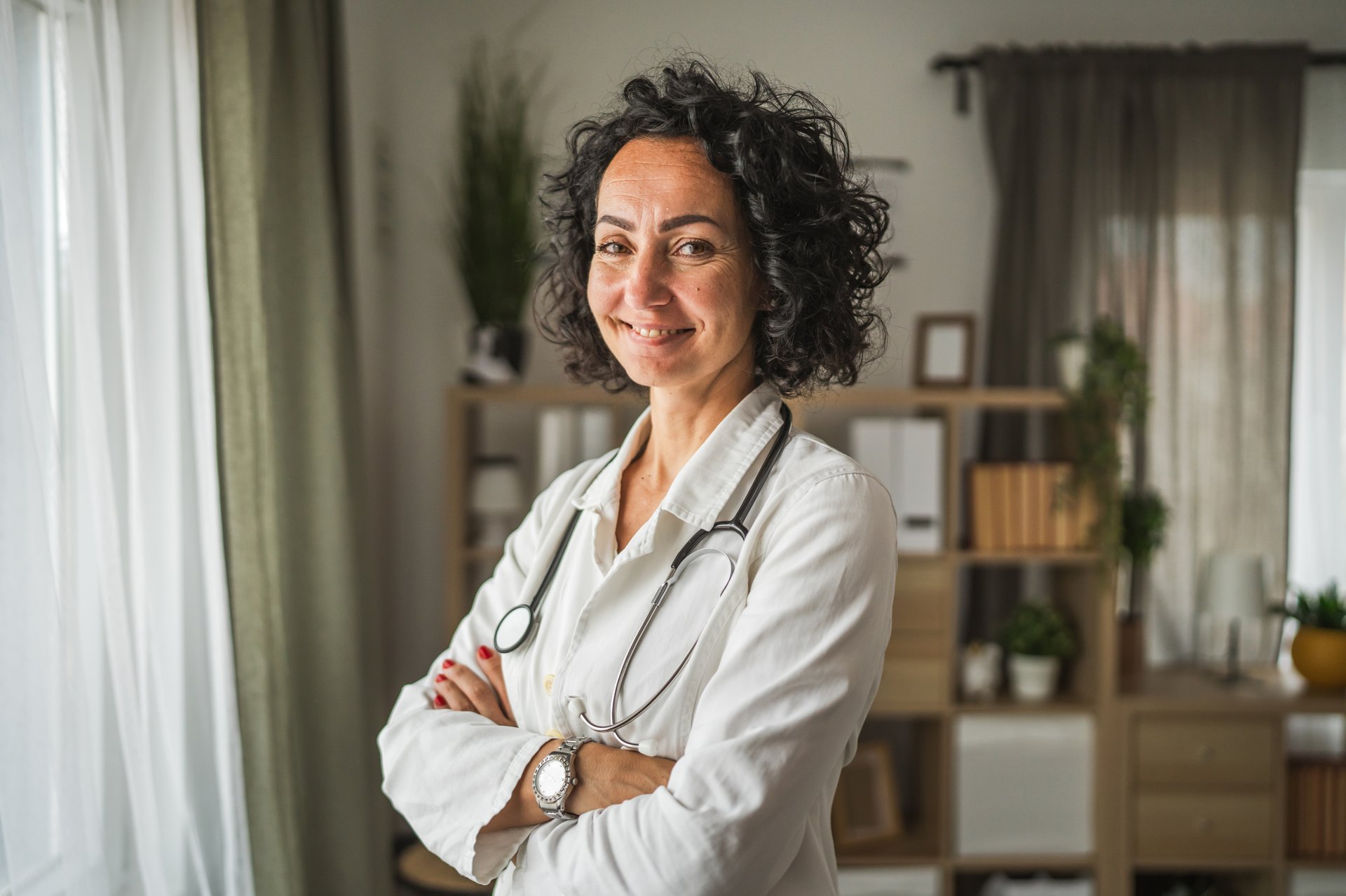 A smiling mature woman doctor with curly hair and a stethoscope stand confidently, look out the window in a bright, modern office with plants and bookshelves