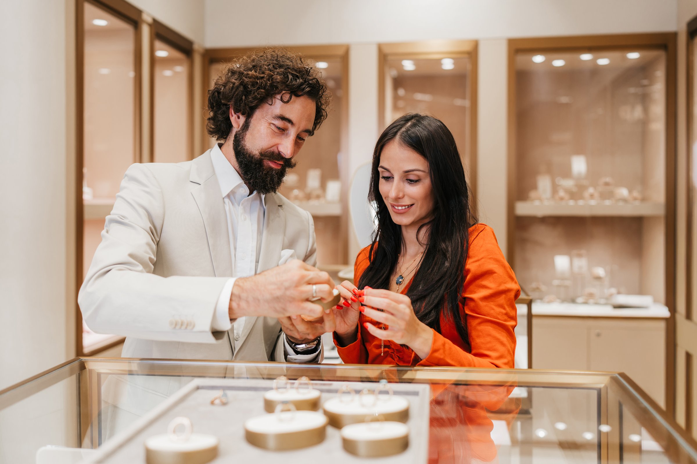 Well-dressed couple at a luxury jewelry boutique examining rings in a glass case, comparing engagement and wedding bands while smiling and preparing for a proposal or special purchase