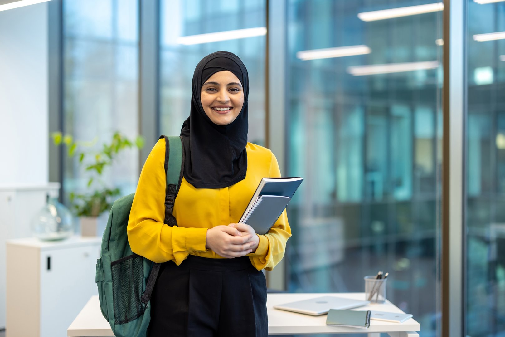 Young Arab student in hijab smiling and looking at camera. Woman inside educational university, holding books and backpack.