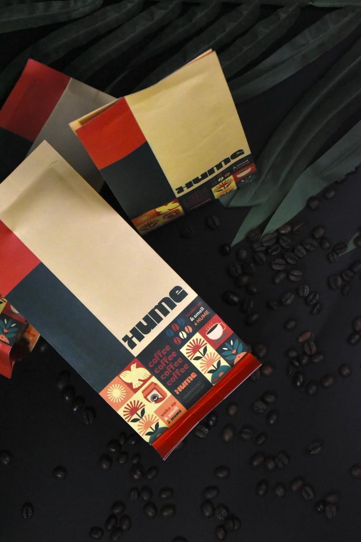 Two bags of coffee labeled 'Hume' with scattered coffee beans on a dark surface, against a leafy green background.