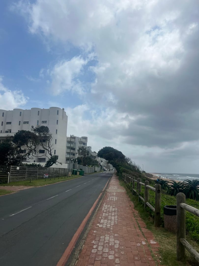 Coastal road with a sidewalk, ocean view on the right, apartment buildings on the left, and cloudy sky overhead.