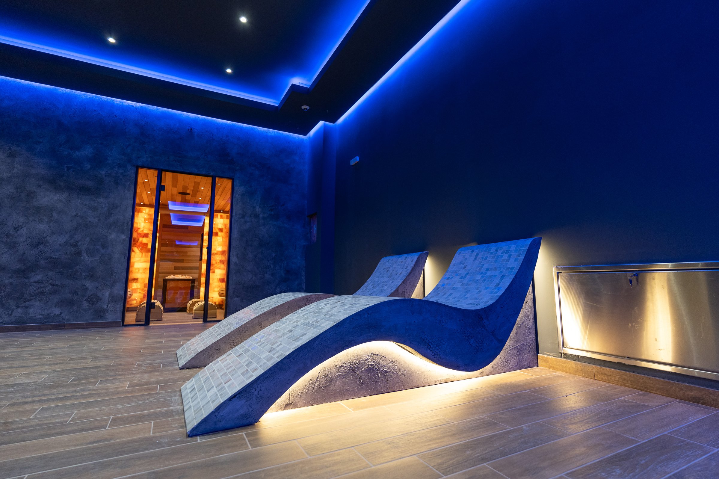 Modern wellness space showcasing elegant heated loungers and sauna entrance, illuminated by soothing ambient blue lighting, evoking calm and relaxation.