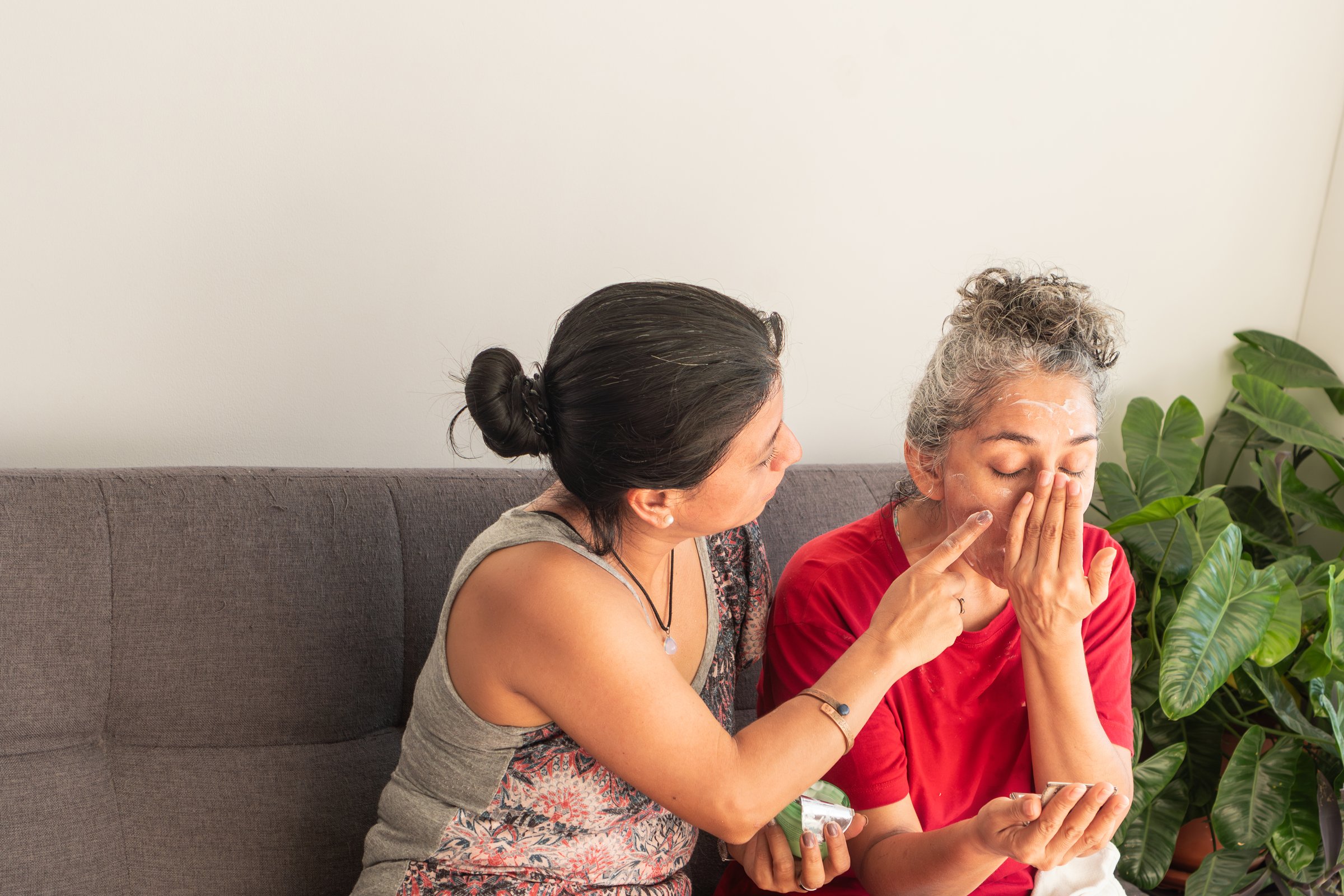 Daughter helping her mother applying moisturizer on her face, sitting on a sofa at home