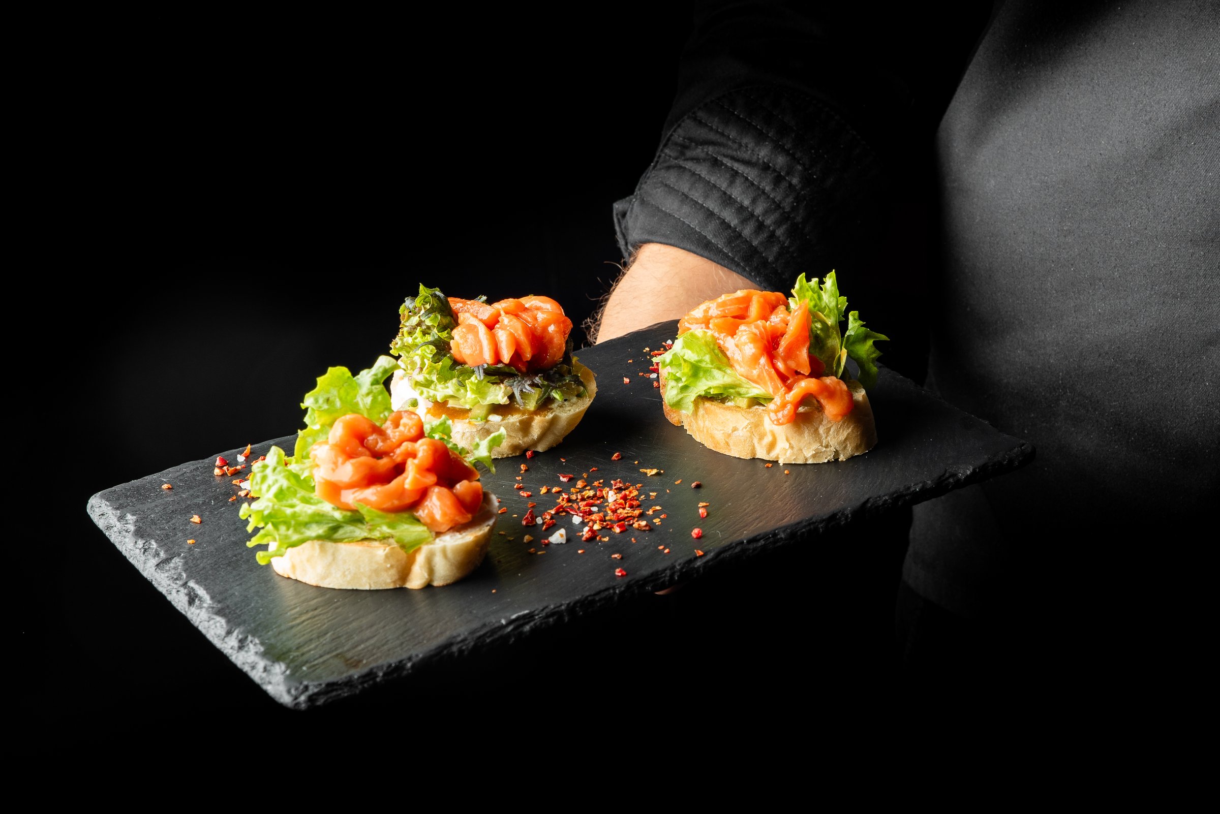 A chef's hand holds a slate platter with three bruschettas topped with fresh lettuce, smoked salmon, and spices. The dark background enhances the gourmet presentation.