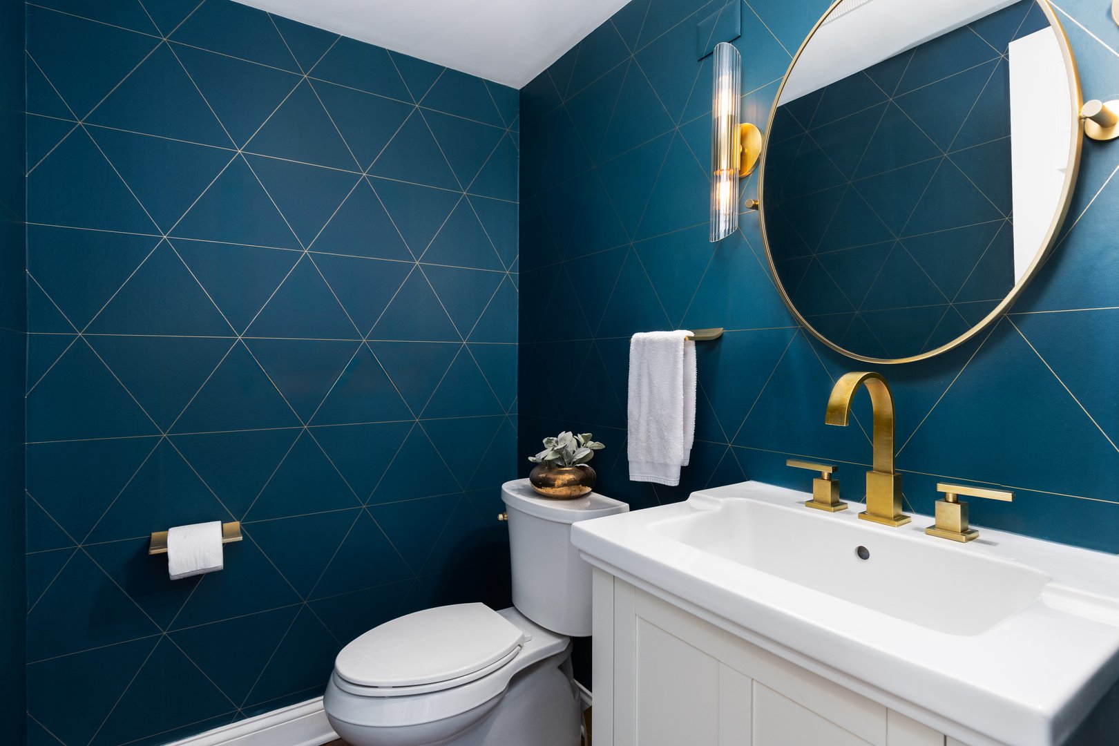 A bathroom with a blue and gold pattern wallpaper, gold faucet and light fixtures, and a white cabinet and sink.