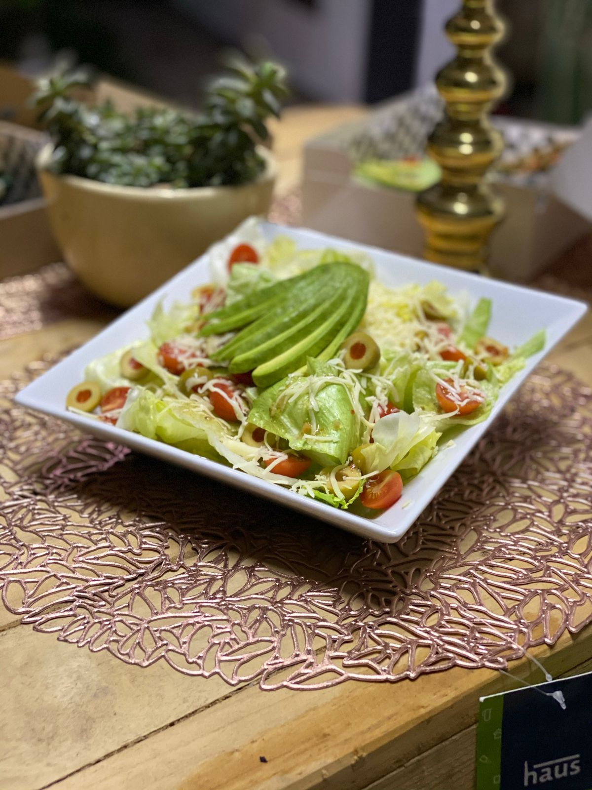 Salad with avocado slices, cherry tomatoes, shredded cheese on a square plate, placed on a decorative table with plants.