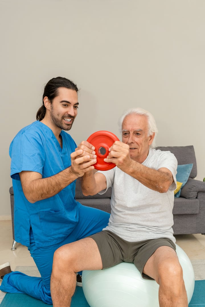 Vertical image of Male Physiotherapist assisting senior man exercising with weights and fitness ball at home