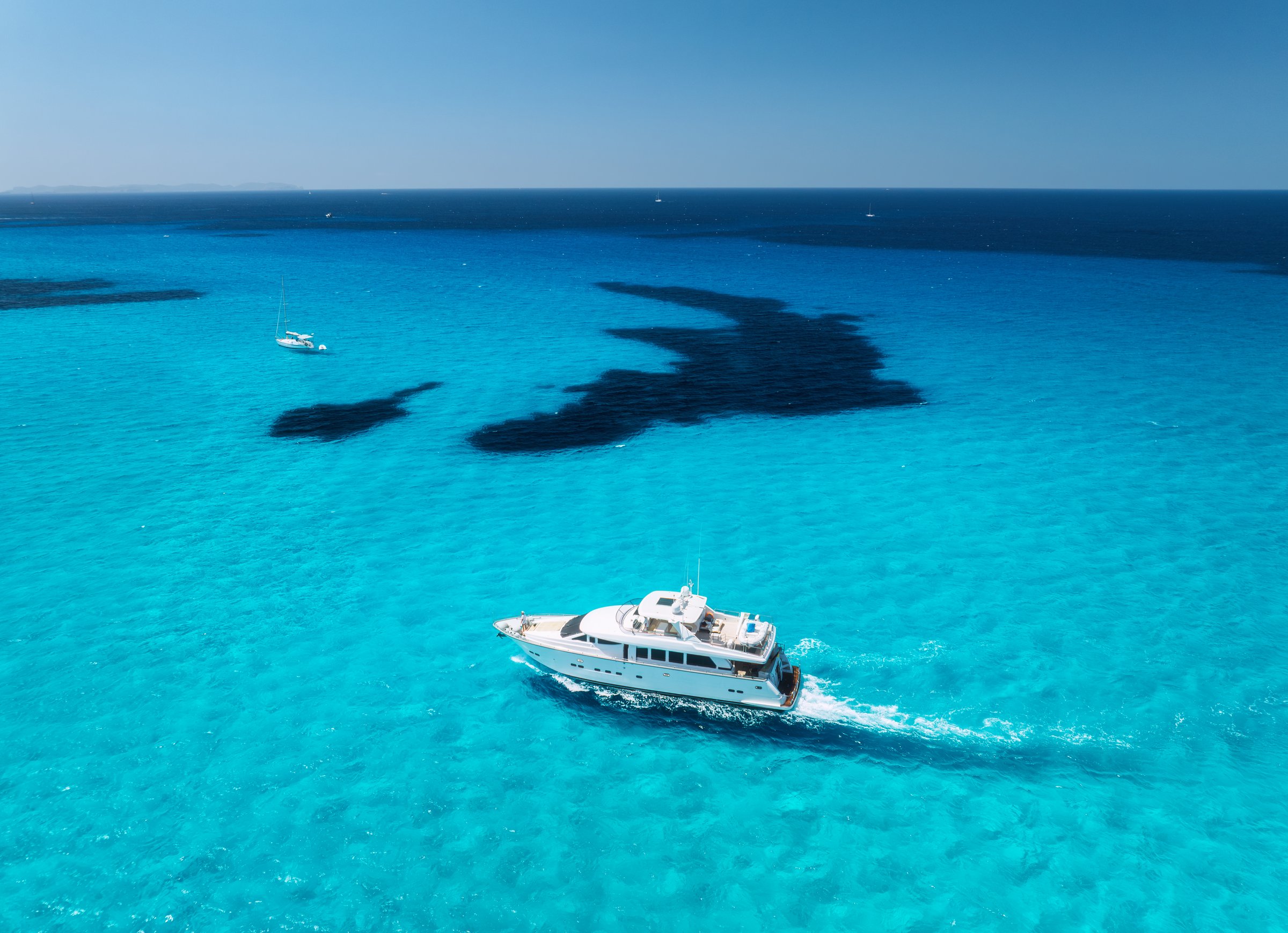 Aerial top view from drone of luxury yacht moving through shallow turquoise sea above dark rocky seabed, sailboats anchored nearby, summer scene near Es Trenc, Mallorca, Balearic Islands, Spain