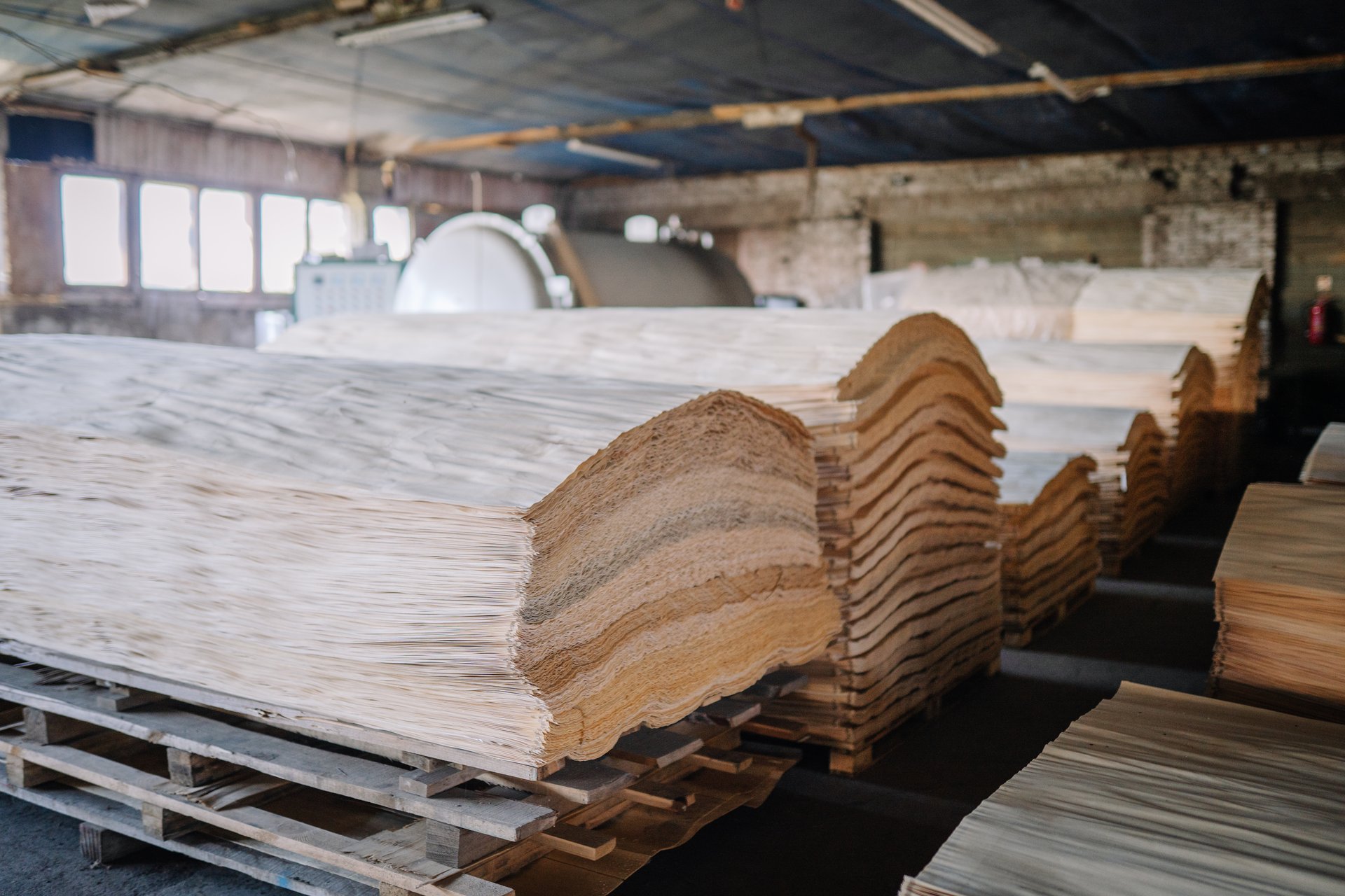 Stacks of sliced wood veneer sheets neatly arranged on pallets inside a woodworking or lumber processing factory with industrial equipment in the background.