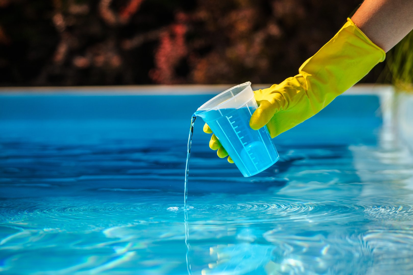 Hand with yellow protective glove pouring blue chemical liquid from measuring cup into swimming pool water for cleaning and disinfection. High quality photo
