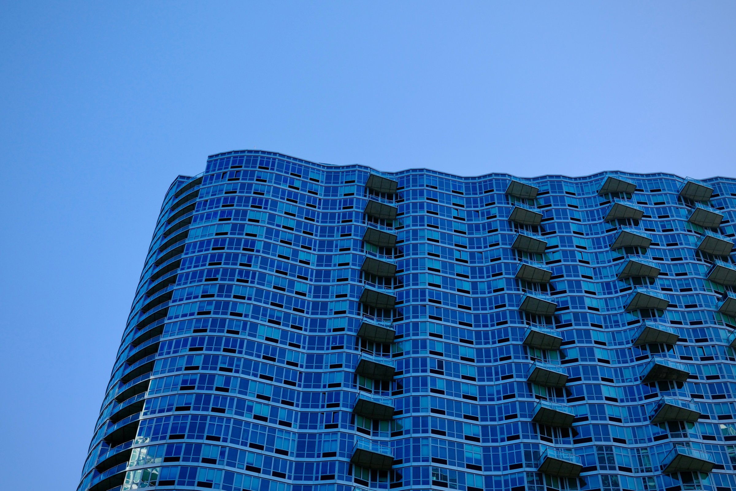 A modern blue-glass residential tower curves in rhythmic waves against a clear sky in Manhattan.