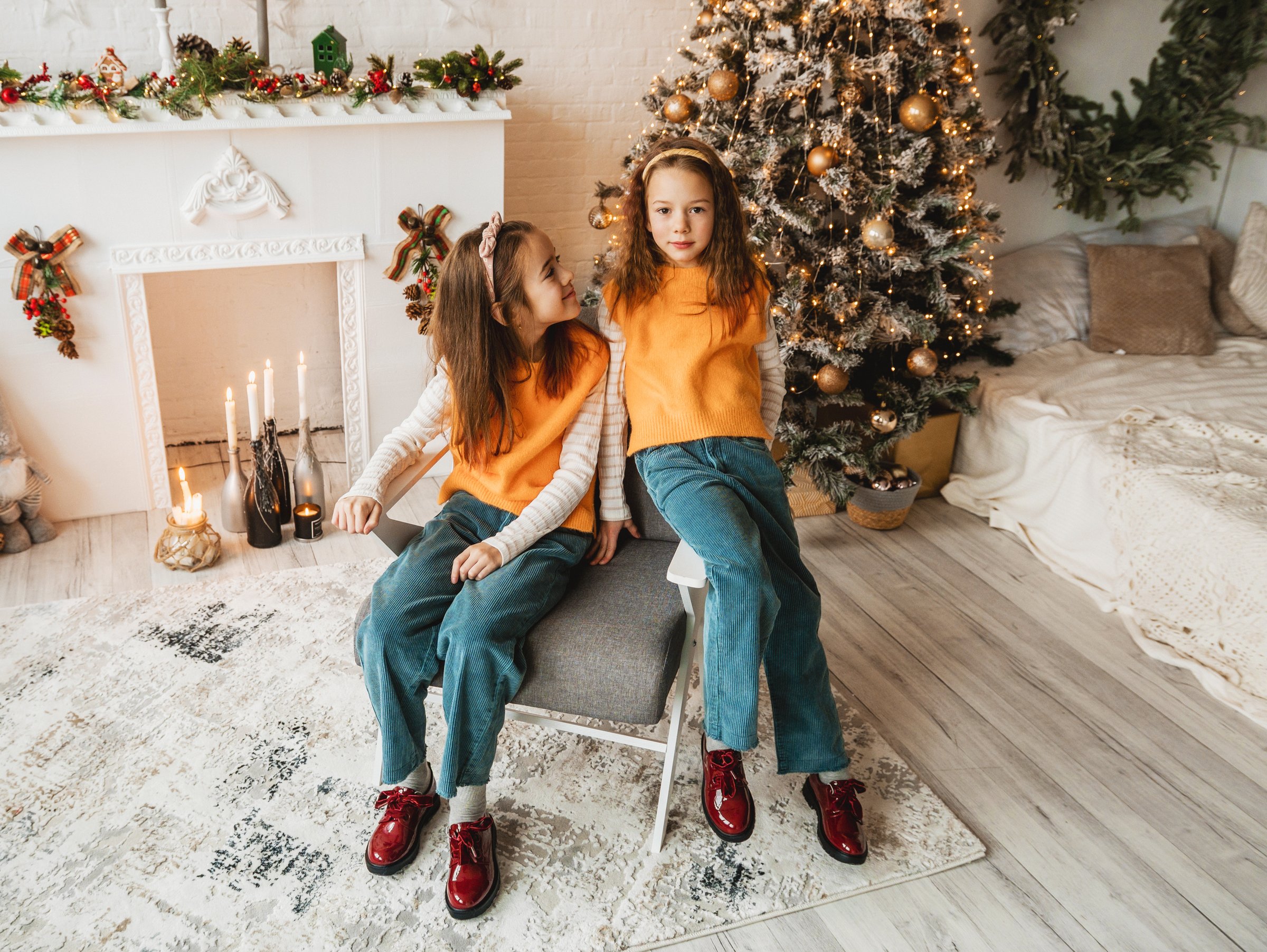 Two adorable sisters in matching outfits pose for a Christmas portrait. They sit near a decorated fireplace and Christmas tree, creating a heartwarming holiday scene.
