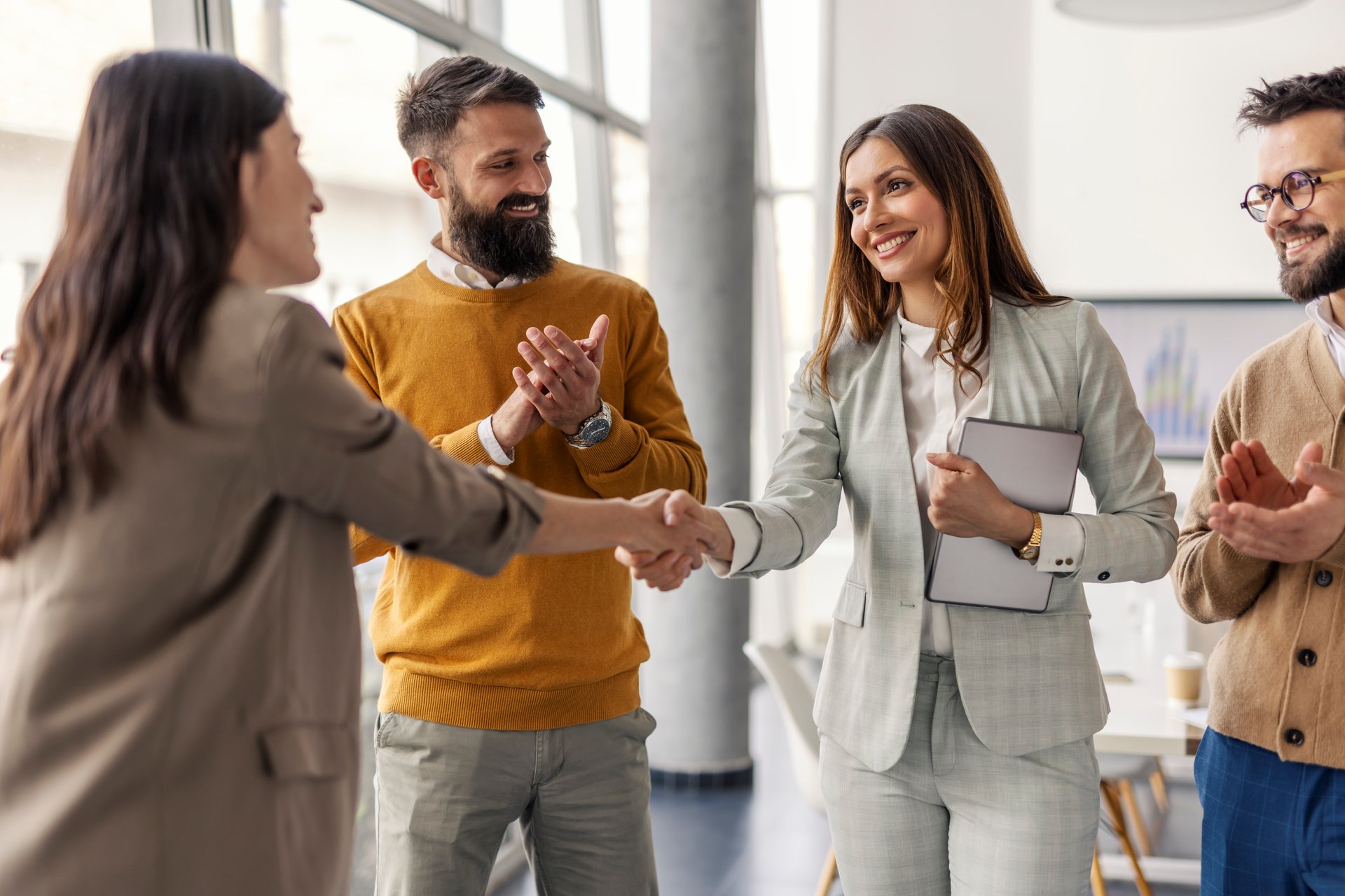 Two successful female executives shaking hands for partnership and standing at office while team members clapping hands at them.