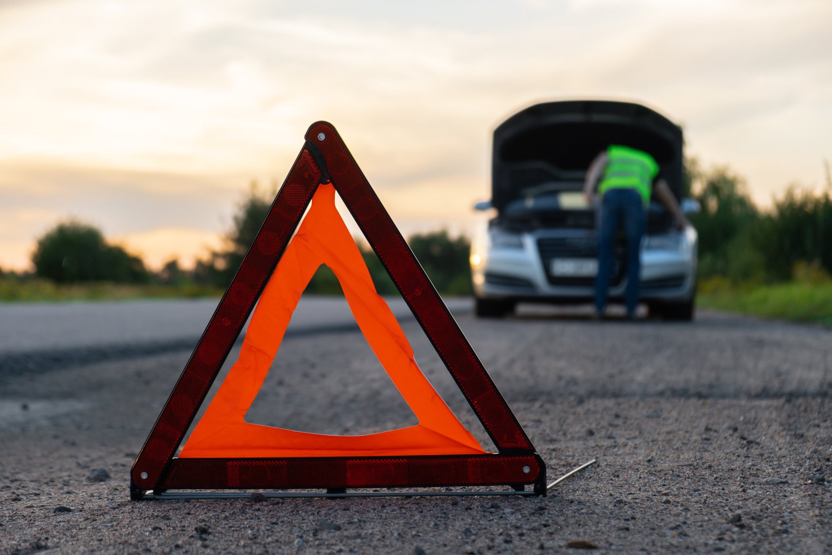 Broken silver luxury car emergency accident. Man driver installing red triangle stop sign on road. Sport automobile turned on blinkers technical problems on the road. Safety procedure vehicle broken down