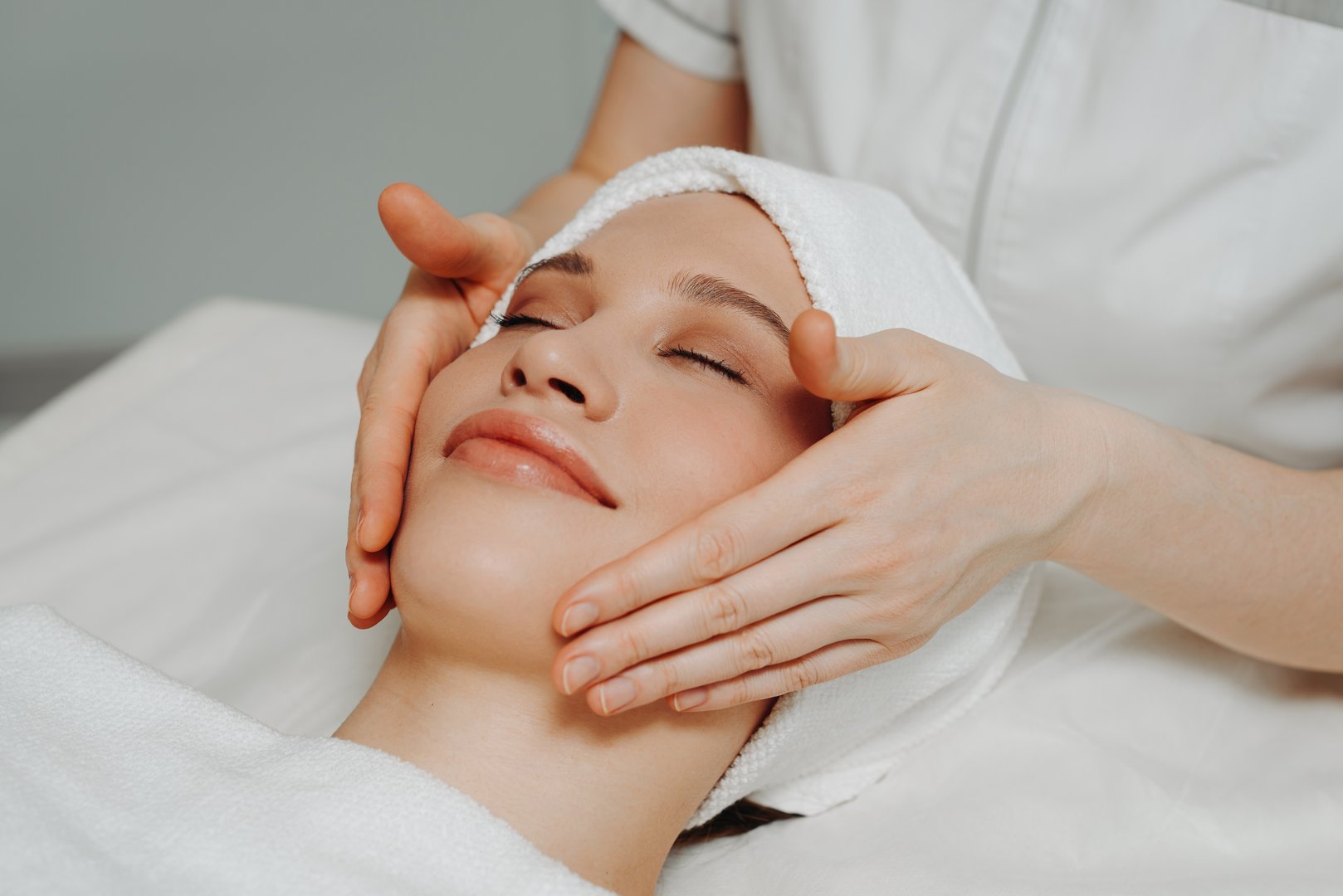 face massage, young attractive caucasian woman, beautiful relaxed lady with closed eyes smiling, white towel, spa therapist doing facial and head massage in beauty salon, skincare and bodycare concept