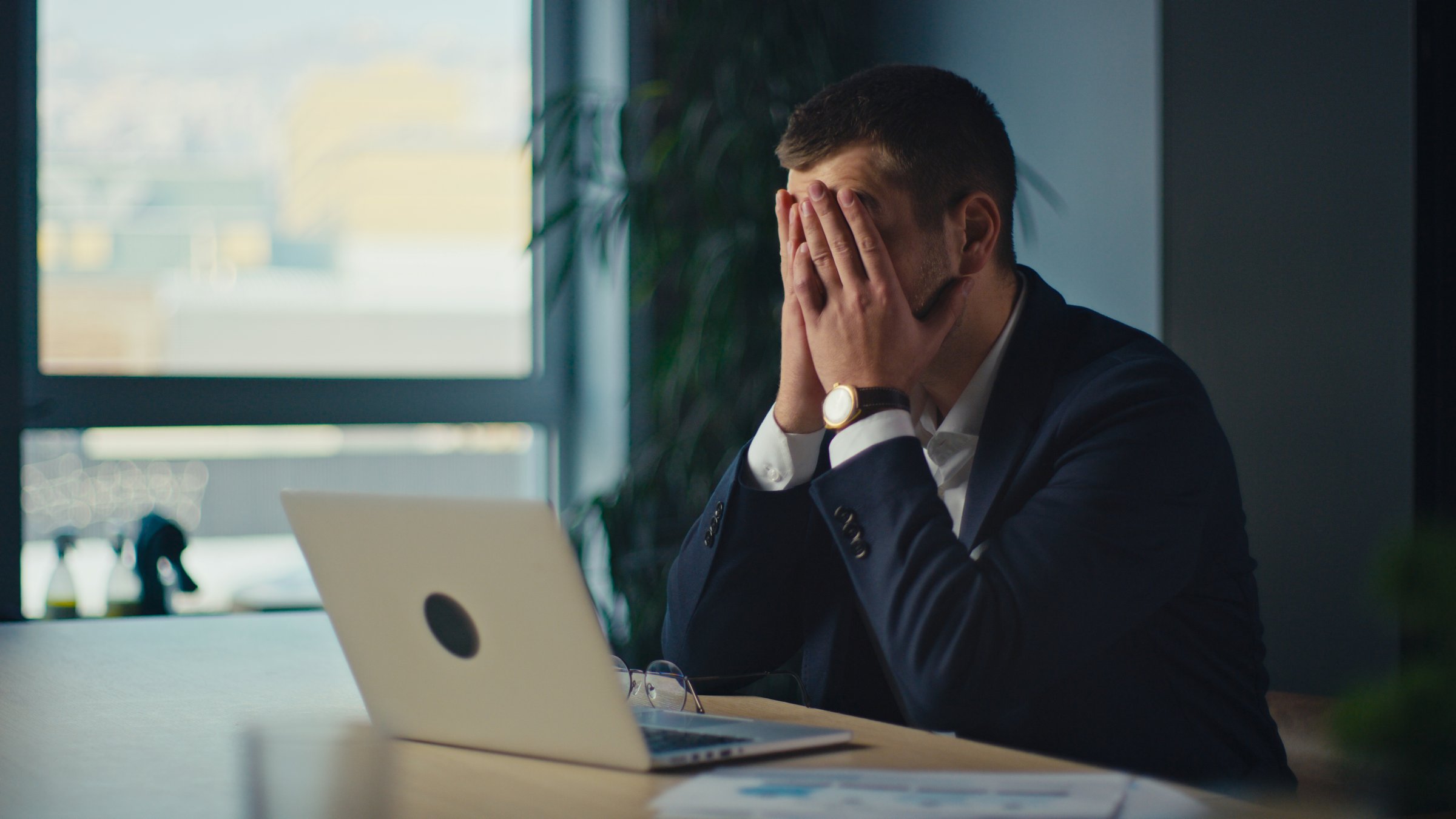 Frustrated businessman in a suit sitting at a desk, covering his face with hands in stress. Modern office environment with a laptop and documents. Business pressure, failure, corporate burnout.