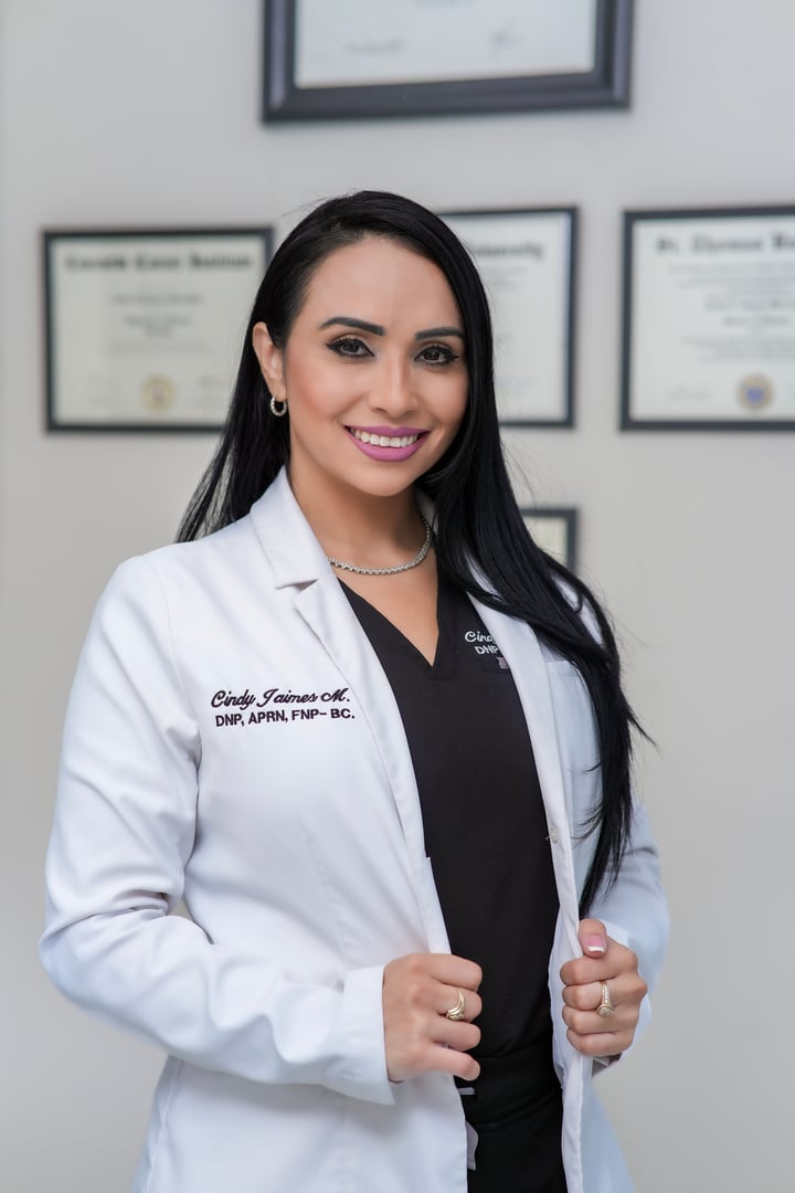 A female medical professional in a white coat smiles, standing in an office with framed certificates on the wall.