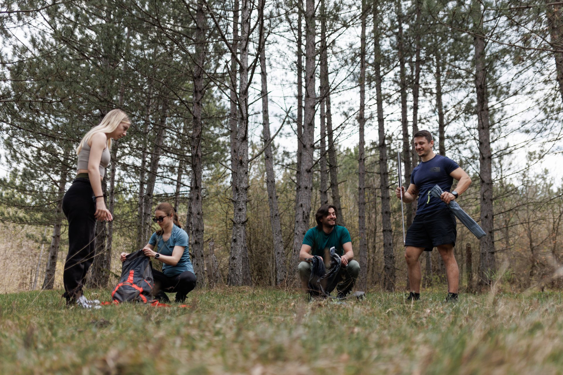 Four friends engage in camping tasks in a green forest clearing, intently organizing gear and enjoying each other's company on a sunny afternoon.