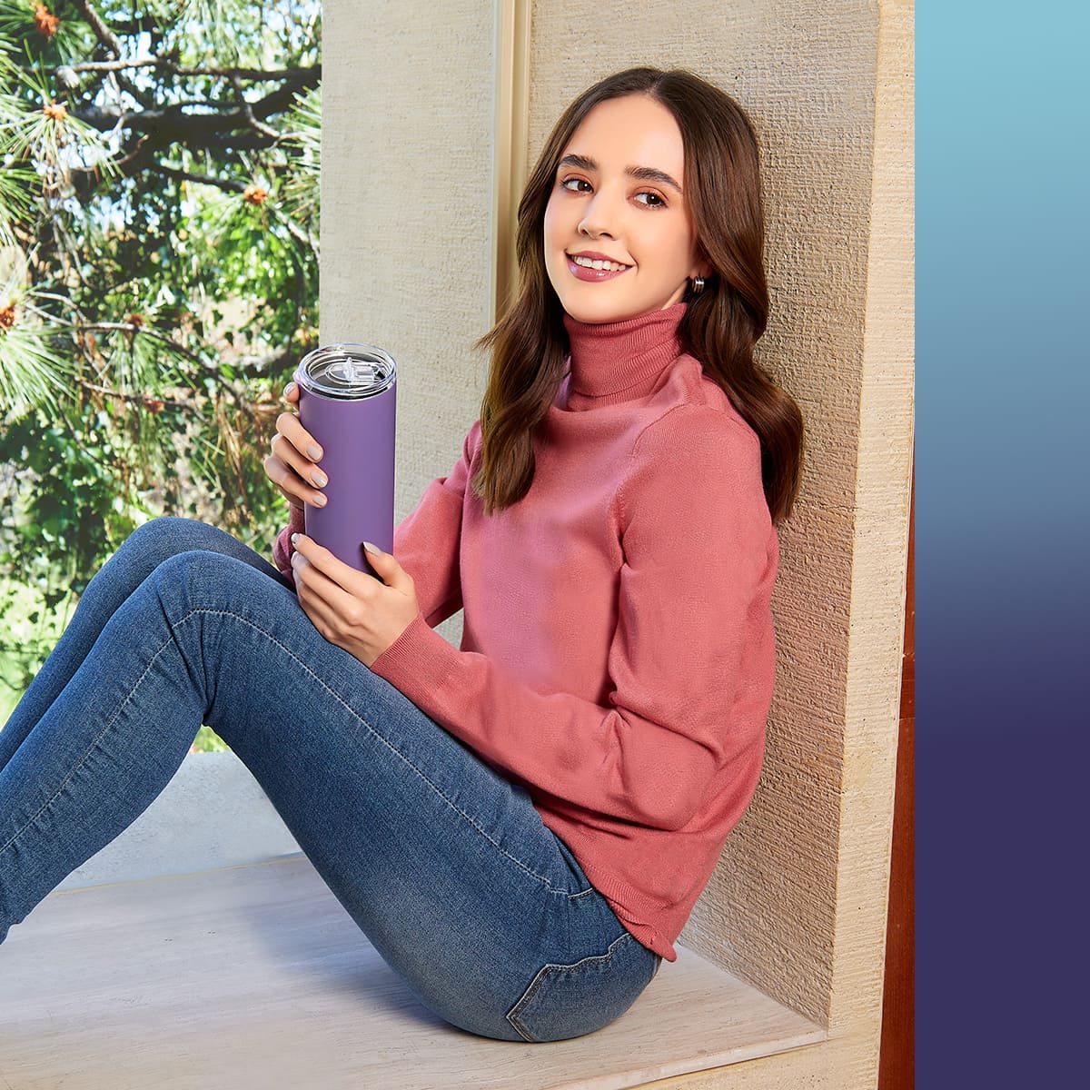 Woman in a pink turtleneck, holding a purple tumbler, seated by a window with trees visible outside.
