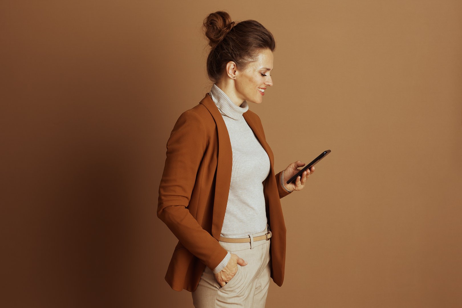 A smiling businesswoman in a brown blazer, standing in profile, casually browses her black smartphone. Her relaxed yet engaged demeanor reflects modern digital communication and efficiency.