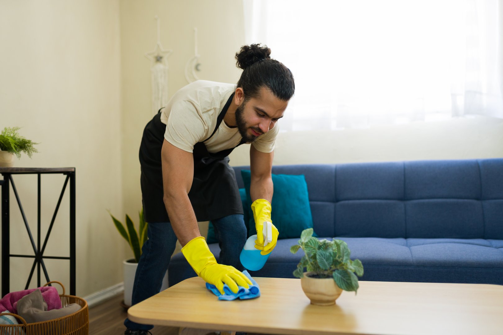 Male housekeeper wearing gloves and an apron diligently cleaning a coffee table in a cozy living room, ensuring a hygienic and tidy space