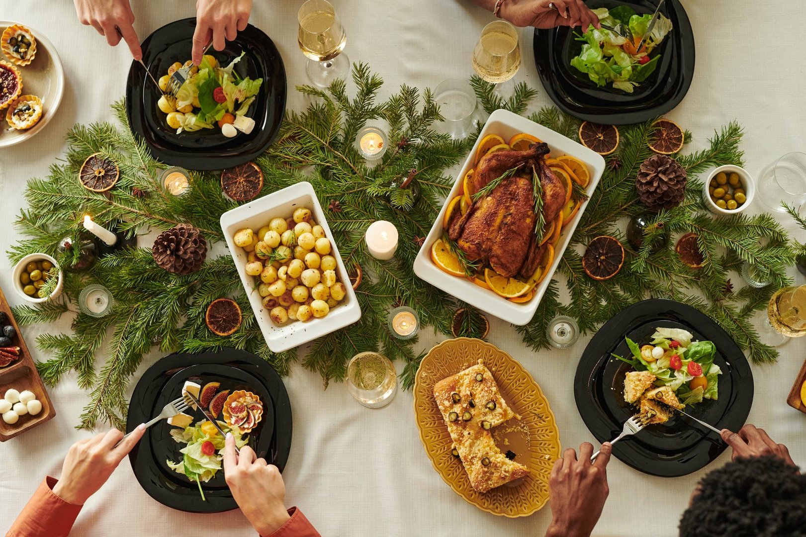 Diverse group of young adults and Young people sharing Christmas meal at decorated table, serving roasted chicken, salad, potatoes, bread, and desserts, hands reaching for food