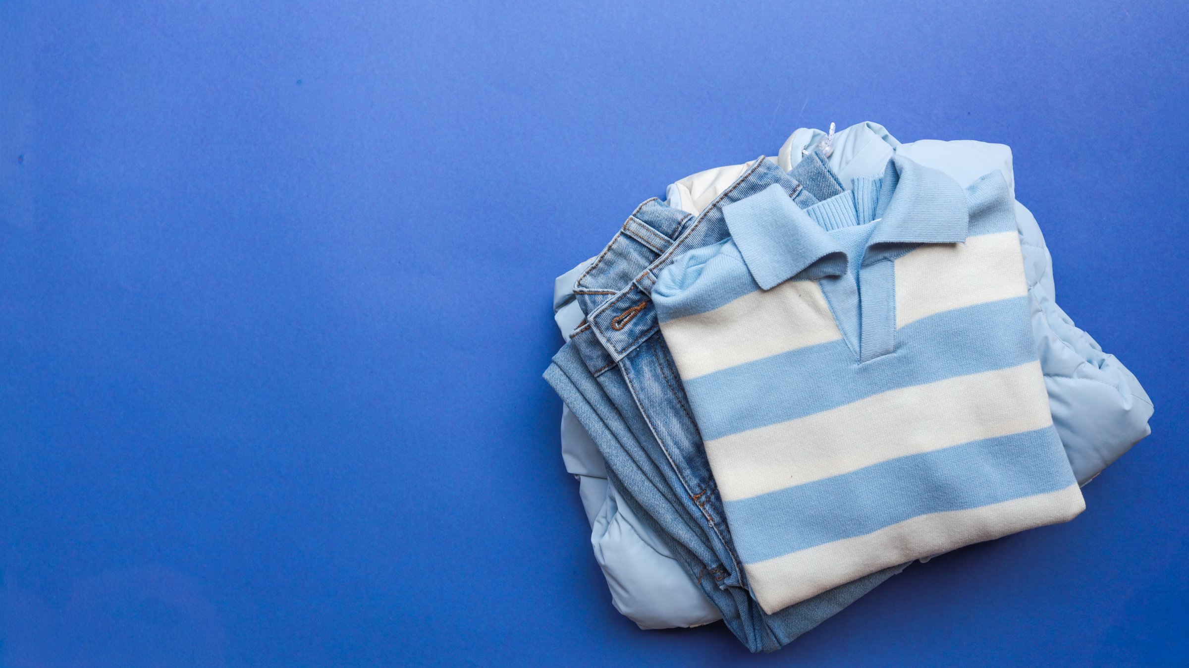 A collection of folded clothing, including a striped polo shirt, jeans, and a jacket, are arranged on a solid blue surface.