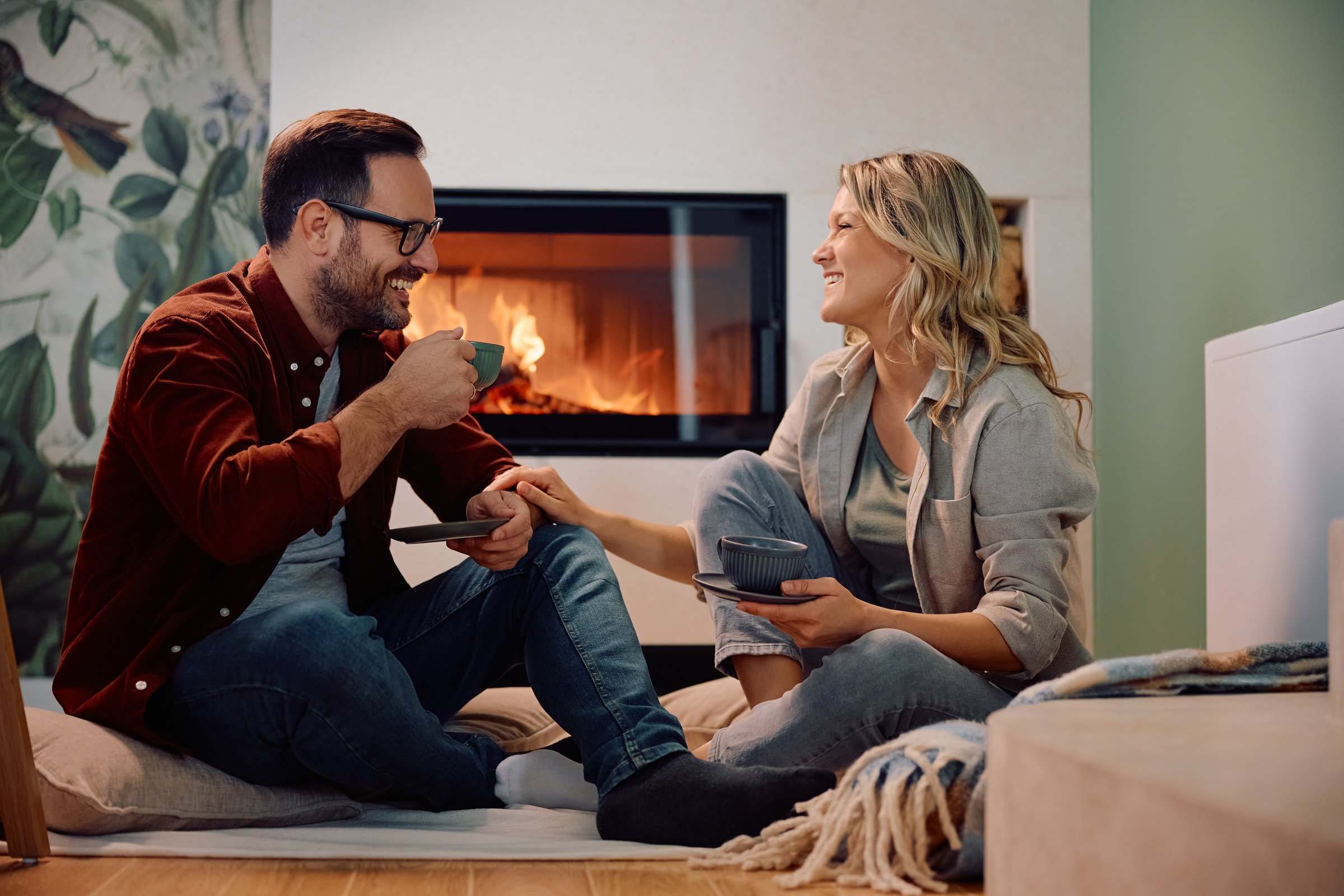Happy couple drinking coffee and communicating while relaxing by fireplace at home.