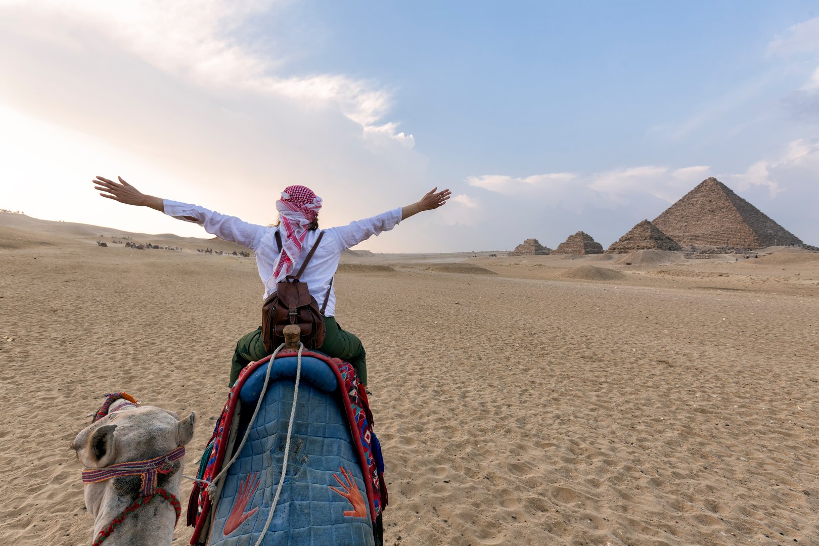 Camels in the Giza Pyramid Complex - A woman with a red turban and arms up riding a camel through thin sand dunes - Cairo, Egypt