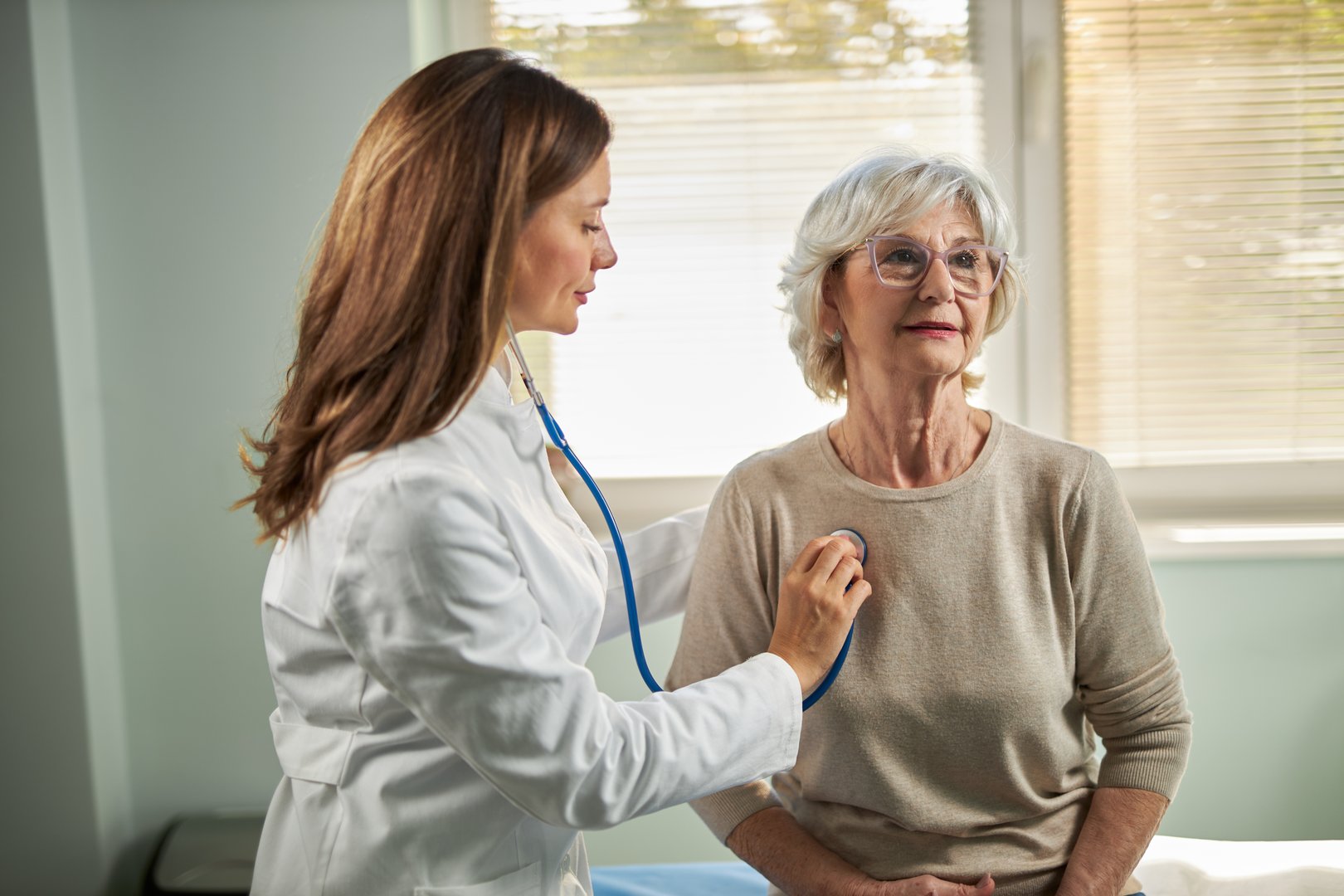 Mid adult female doctor uses a stethoscope to check the heart and lungs on a senior female patient.