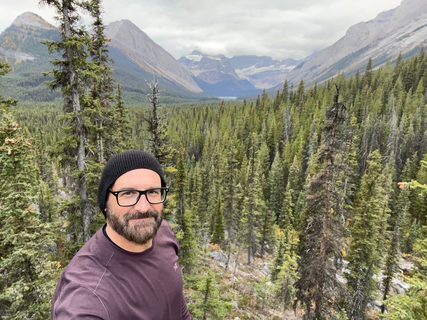 Man wearing glasses and a beanie stands in a forested mountain landscape with cloudy skies in the background.