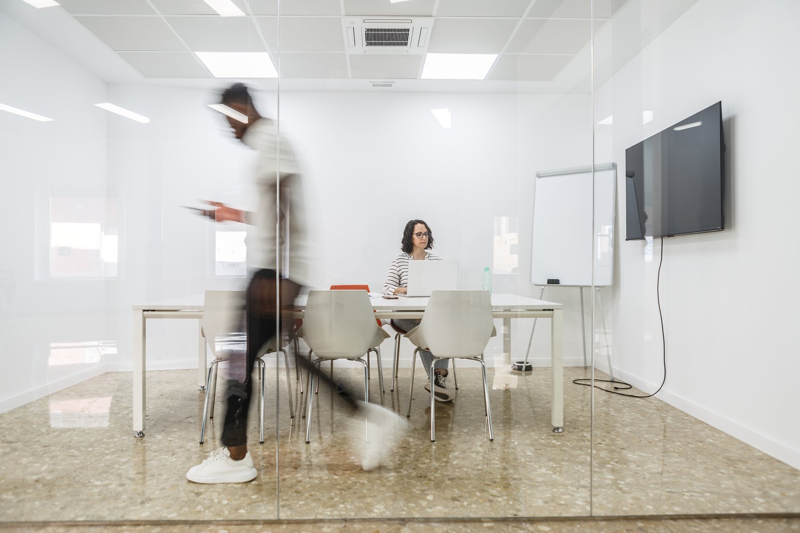 A man in a white shirt walks through a conference room with glass walls, his form blurred in motion. A woman sits at a table with a laptop, working in the room.