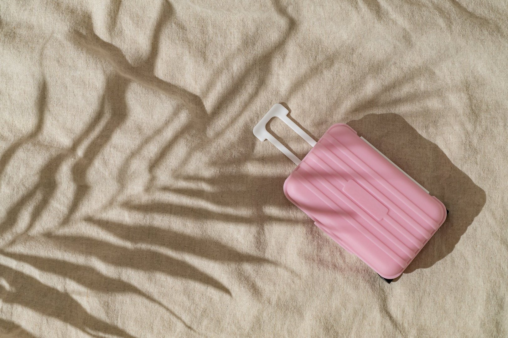 Summer day scene with tropical palm tree leaf shadow and pastel pink baggage on sand color linen background. Minimal stylish travel concept. Trendy summertime adventure idea. Flat lay, top view.