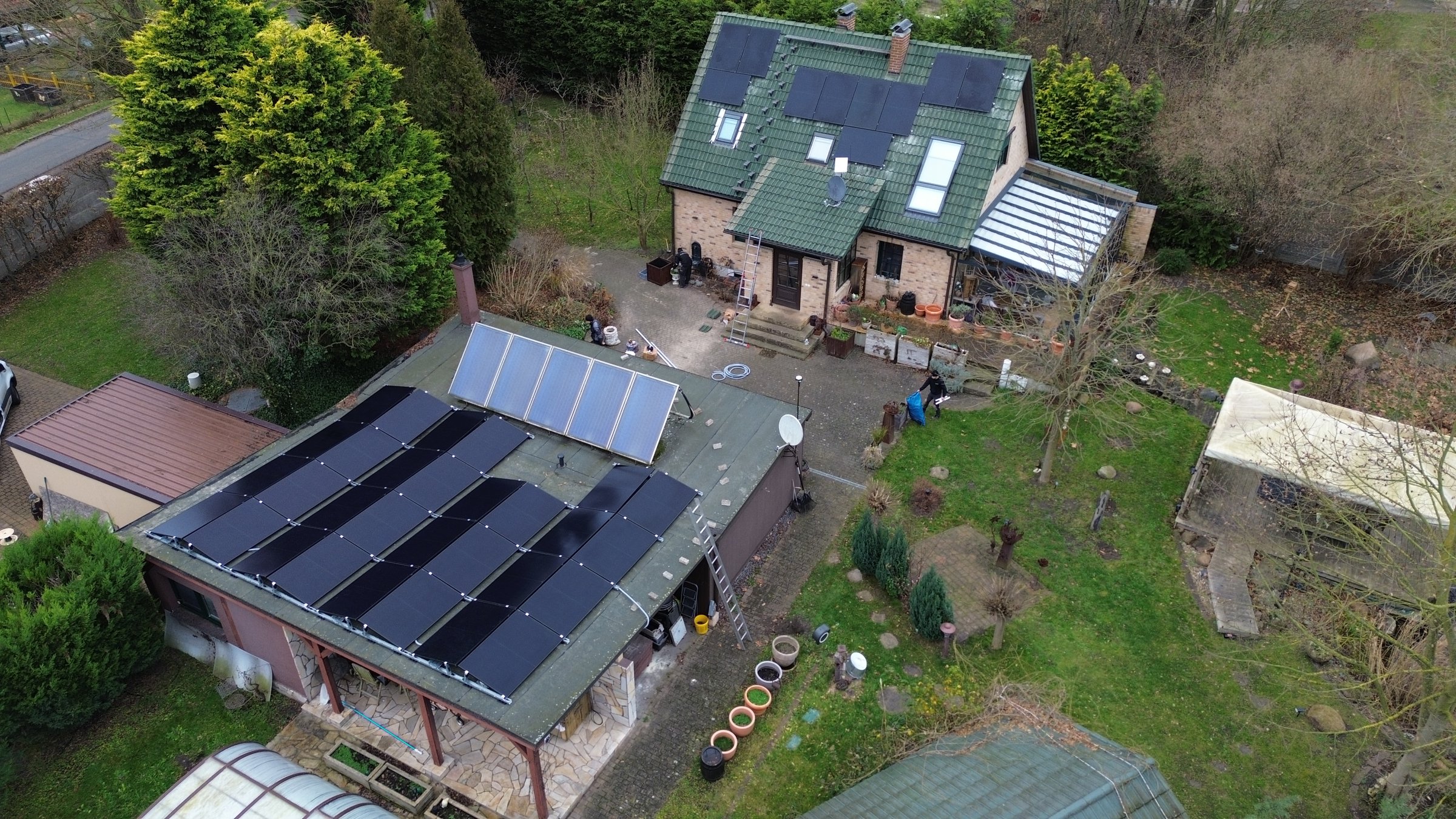 Aerial view of a house and a building with solar panels on the roofs, surrounded by trees and garden area.