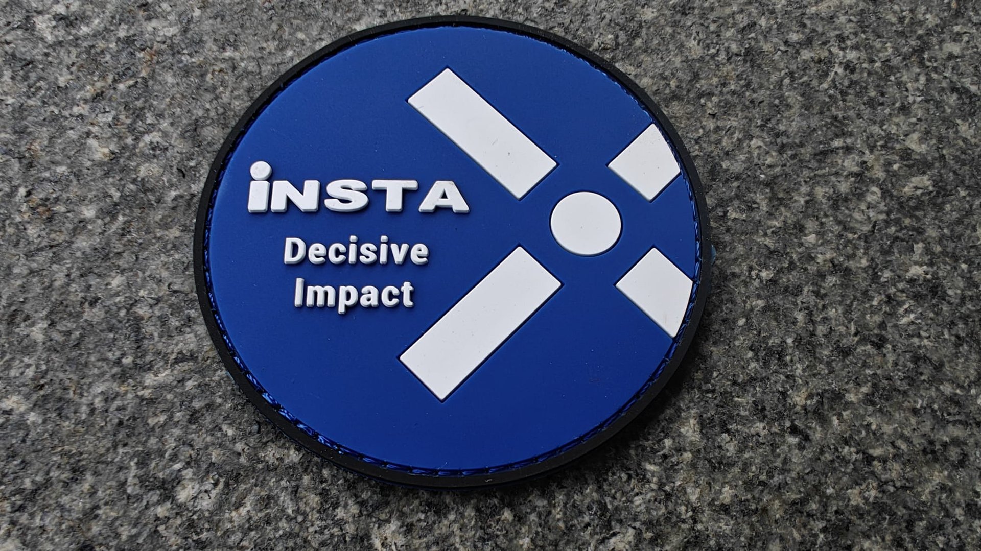 Round blue patch with the text Insta Decisive Impact and white geometric design on a gray textured background.