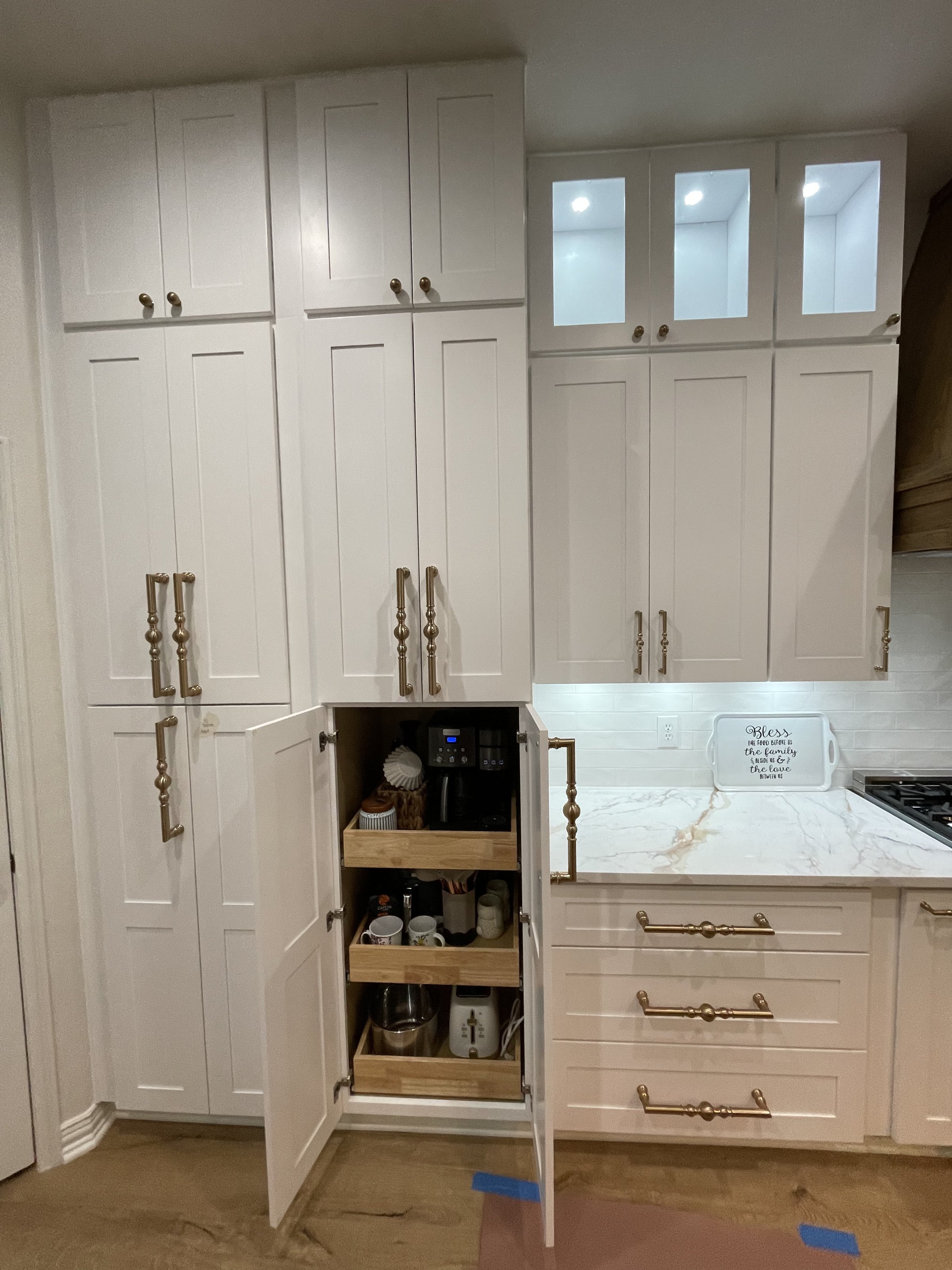 White kitchen cabinets with gold handles, open to reveal pantry shelves filled with jars and a coffee machine.