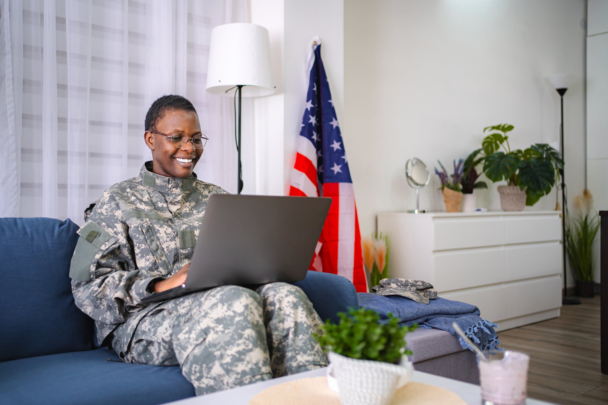 Smiling mid adult african american female soldier in uniform working on a laptop at home with United States flag hanging on the wall