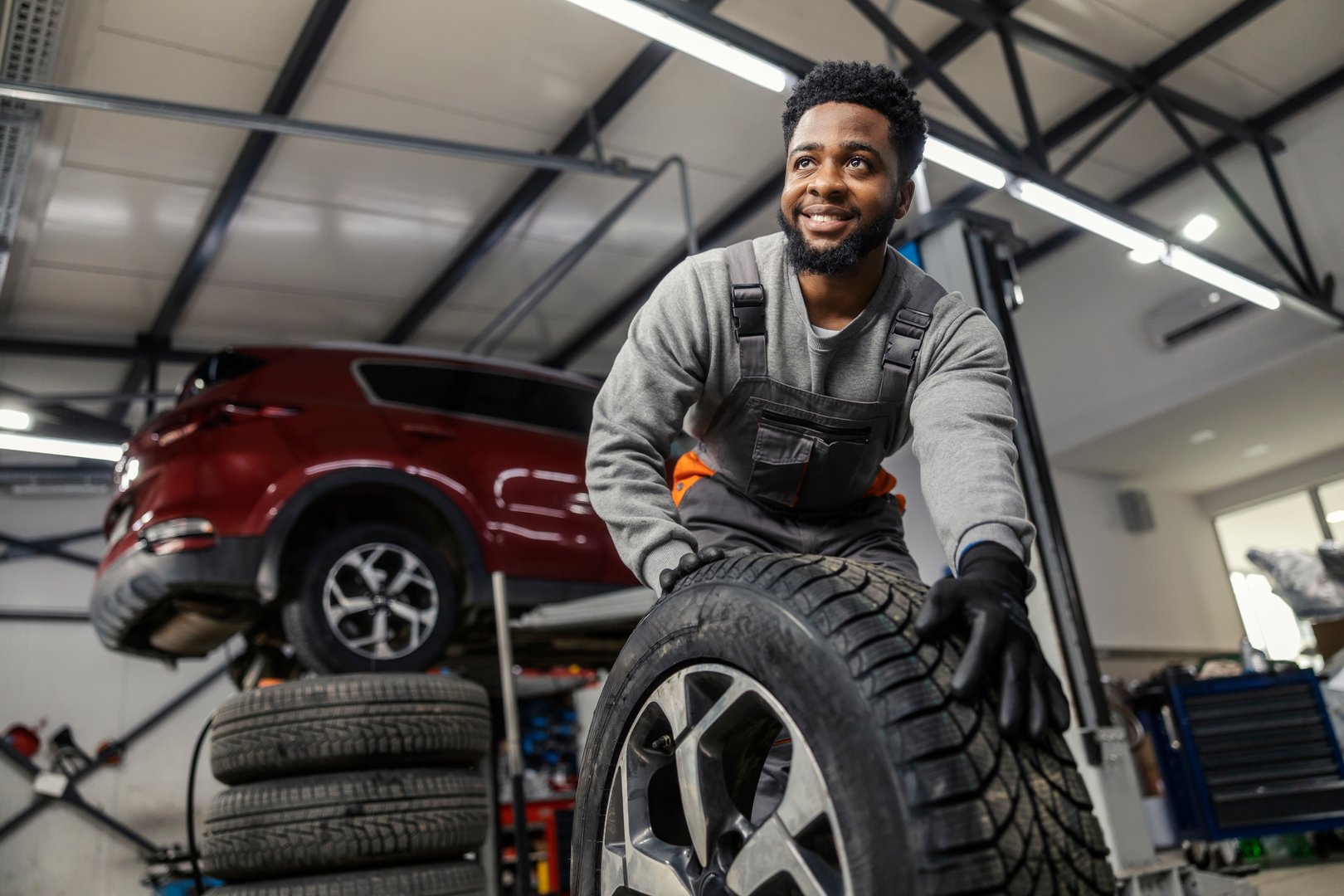Portrait of hardworking smiling car technician rolling car tyre towards camera at automotive service center.
